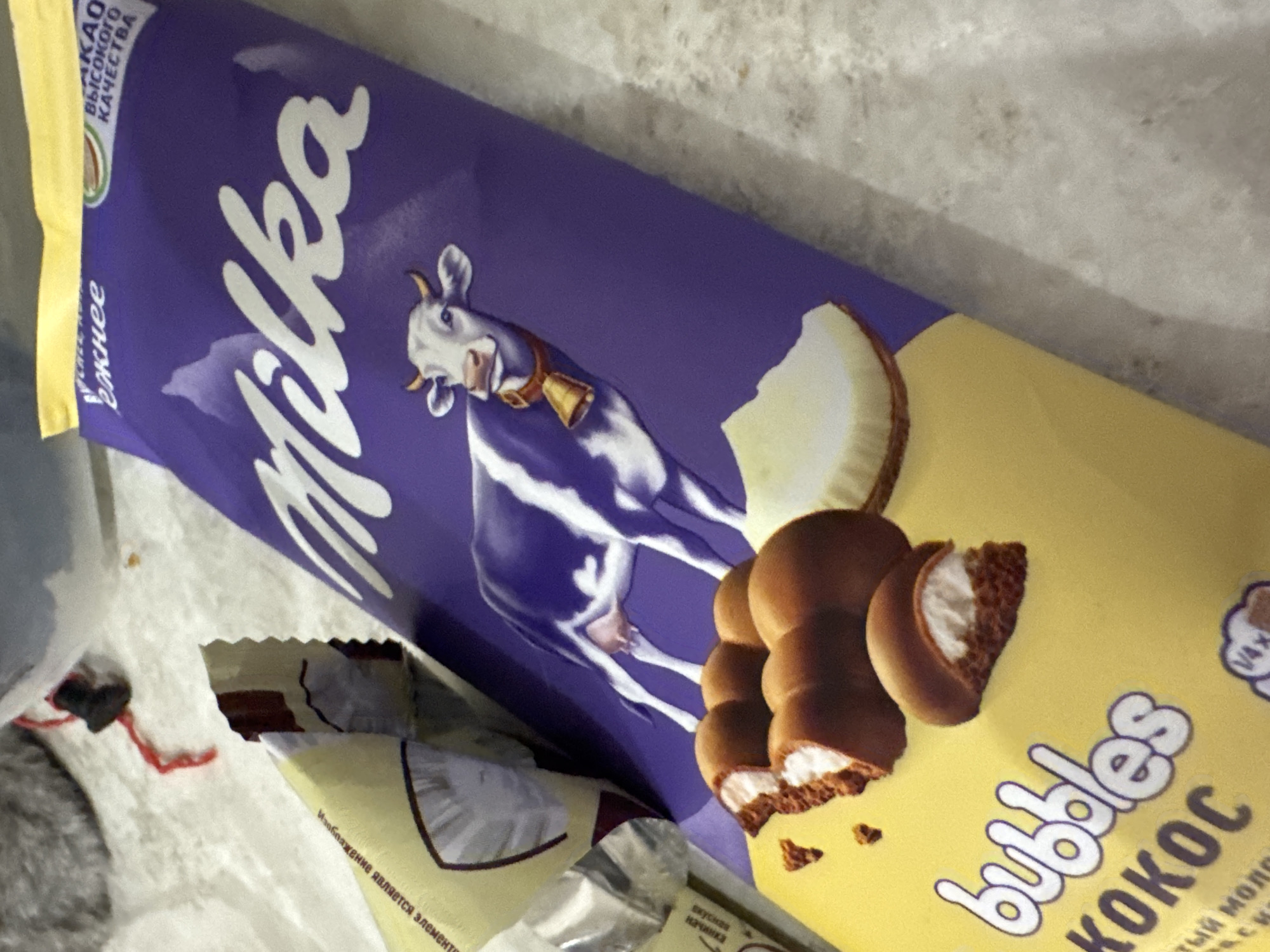 The image shows a close-up of a Milka "Bubbles Kokos" chocolate bar. The packaging is predominantly purple with a yellow section at the bottom. The Milka logo is prominently displayed in white lettering across the purple section, above a depiction of a cow wearing a bell. A portion of the chocolate bar is visible at the bottom, showing several pieces of chocolate with a white, creamy filling. There are also some crumbs scattered around the chocolate pieces. Part of the wrapper is torn open, revealing more of the chocolate inside. The background appears to be a textured surface, possibly a countertop. The scene is not identifiable as being in the city of Saratov, Russia.