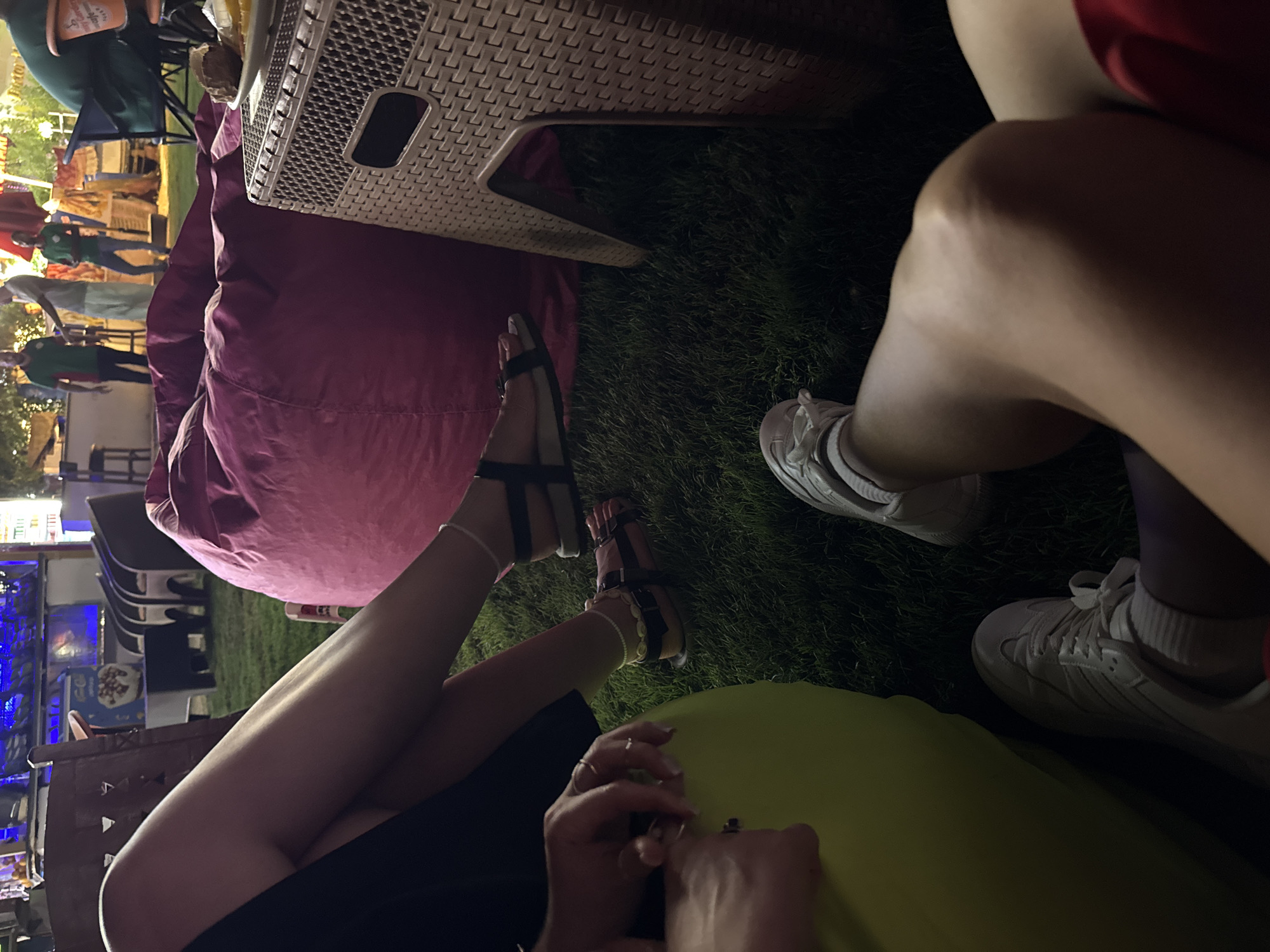 The image depicts a group of individuals relaxing on beanbag chairs in an outdoor, dimly lit setting. The ground is covered in grass, and in the background, a lively scene unfolds with people standing and a bar-like structure with bright lights. One person is wearing a black dress, another a red top, and there are glimpses of legs in white sneakers and sandals. A pink beanbag chair is prominent in the foreground, alongside a woven-texture side table with a plate of food. The overall impression is a casual, social gathering. The location is identified as Unknown, Egypt.