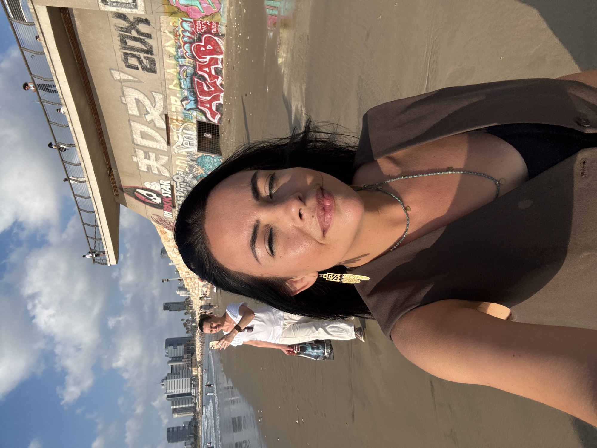 This is a selfie taken on the beach in Tel Aviv, Israel. The foreground shows a woman's face, with her eyes mostly closed, looking towards the camera. She is wearing a dark vest over a black top and a multi-layered necklace. To her left, further down the beach, another woman is holding up a smartphone to take a photo. She is wearing a white t-shirt and light-colored pants, and carrying a shopping bag. The background shows a section of the Tel Aviv coastline with buildings in the distance and a walkway with graffiti on the wall below it. The sky is blue with some clouds.