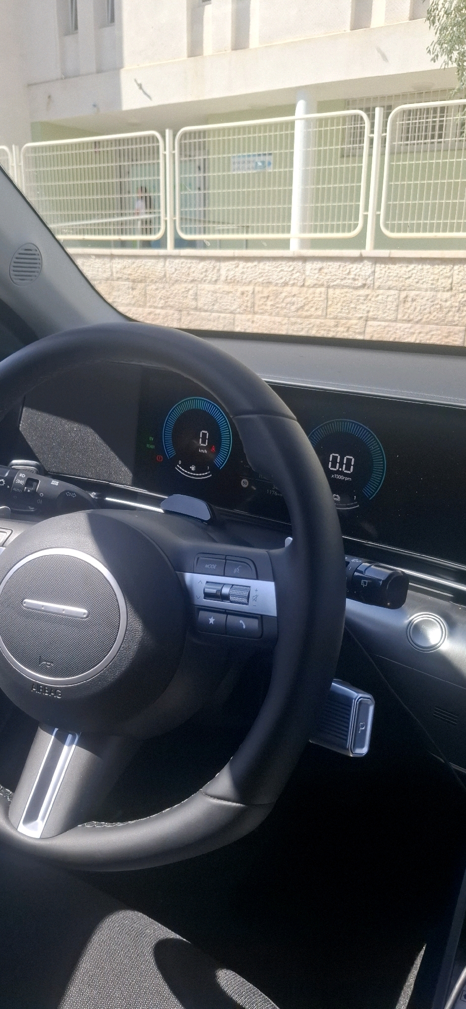 A first-person perspective image captures the interior of a car, likely a Hyundai Ioniq 5, as evidenced by the digital dashboard displays and steering wheel design. The digital dashboard shows a speed of 0 km/h and an engine speed of 0 rpm, indicating the car is stationary. The exterior view, seen through the car's windshield, reveals a building with light-colored walls and windows, characteristic of Israeli architecture. In the background, a light-colored metal fence with horizontal bars encloses a green area, likely a schoolyard or park, and two figures, possibly children, are visible behind the fence, partially obscured. This scene is consistent with the environment found in Dimona, Israel, which has a prominent educational sector and distinct architectural styles. The car's interior is modern, with a black leather steering wheel, silver accents on the controls, and a digital instrument cluster. The ambient lighting suggests it is daytime, with sunlight casting shadows on the car's interior and highlighting the textures of the dashboard and seats.
