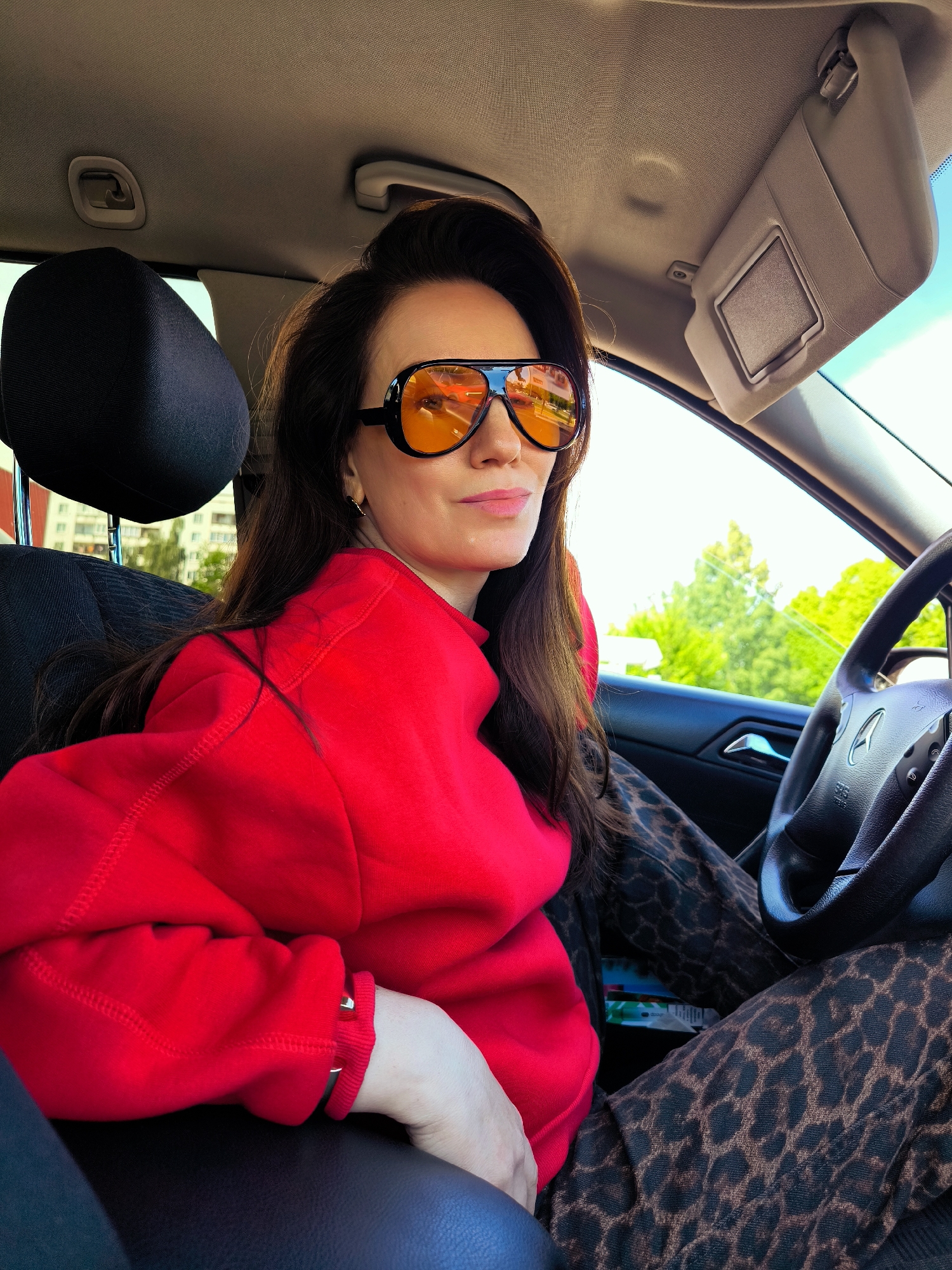 A woman with long, dark hair is seated in the driver's seat of a Mercedes-Benz, possibly parked or stationary. She is wearing large, black-framed sunglasses with orange lenses, a bright red sweatshirt, and leopard-print pants. The interior of the car is visible, including the steering wheel, dashboard, and sun visor. Outside the car, trees and buildings are visible through the windshield. The location appears to be Riga, Latvia, based on the architecture of the building visible through the windshield, and some visible outdoor features that point to an Eastern European/Baltic architecture style.