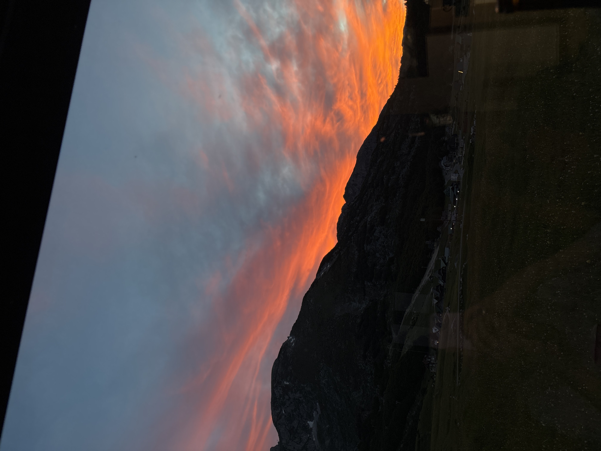 The image shows a mountain landscape at dusk, likely near Virak, Montenegro. The sky is a vibrant mix of orange, pink, and blue, with streaks of clouds. A dark, silhouetted mountain range dominates the left side of the frame, with some snow patches visible on its slopes. In the foreground, a grassy field is visible, with scattered lights indicating a small settlement or village nestled at the base of the mountains. The scene appears to be viewed through a window, with reflections on the glass. There are no people or animals clearly visible in the image.