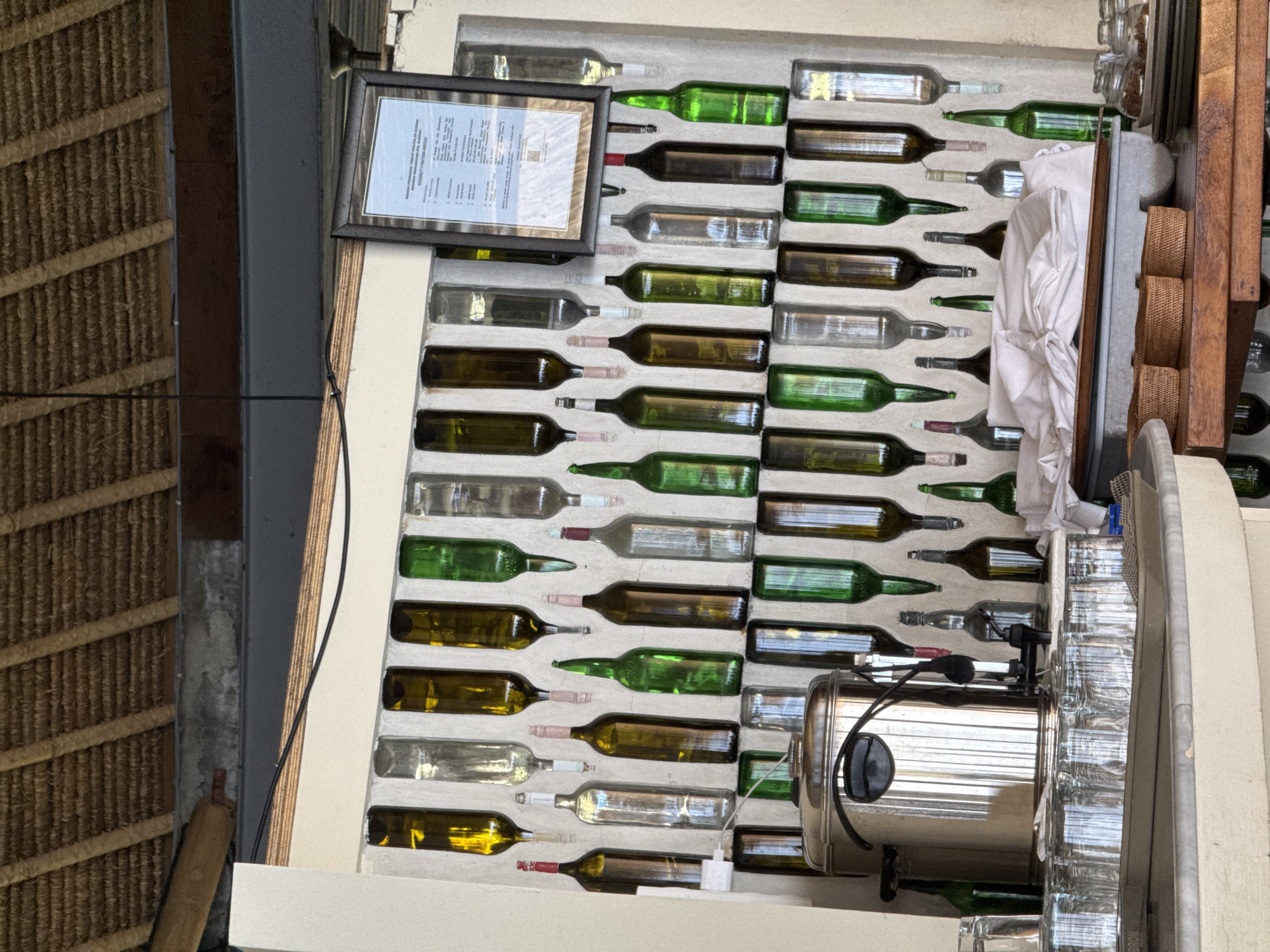 This image depicts a wall decorated with an assortment of empty glass bottles, primarily wine bottles of various colors including green, clear, and brown. The bottles are arranged in staggered rows, with some upside down and others right-side up, suspended from the wall. A framed document, possibly a certificate or notice, hangs on the wall to the right of the bottles. Below the bottle display, a stainless steel beverage dispenser is visible, flanked by stacks of clear drinking glasses. A tray filled with these glasses sits on a marble countertop. The background shows a thatched roof structure, indicative of a tropical or resort-like setting. The scene appears to be in Manggis, Indonesia, likely a restaurant or bar area given the presence of glassware and a beverage dispenser. There are no people visible in the image.