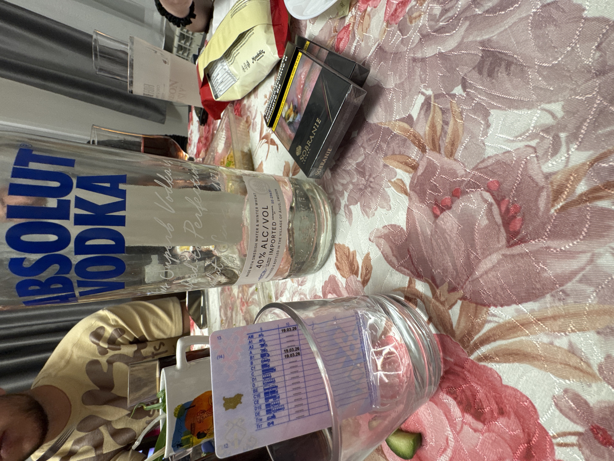 A close-up, eye-level shot depicts a table setting featuring several items suggestive of an informal gathering. On a floral patterned tablecloth are a bottle of Absolut Vodka, a glass containing what appears to be a driver's license, a pack of Sobranie cigarettes, and a bag of Mondelez snacks. The driver's license in the glass has "19.03.26" printed on it. The location in the city of Topoli, Bulgaria cannot be determined from the image. A person wearing a camouflage shirt is partially visible at the bottom left of the frame, seemingly holding a mug with artwork.