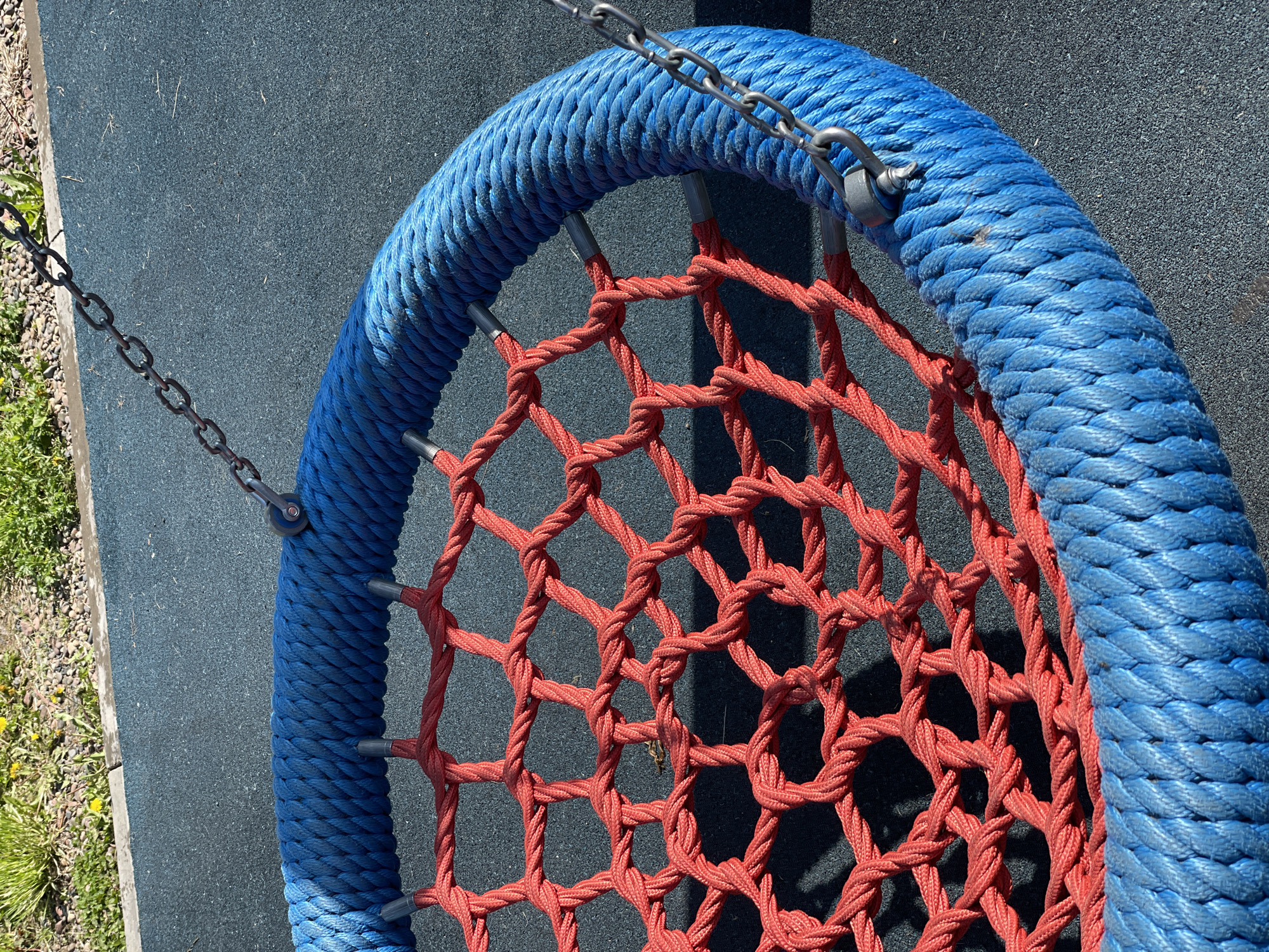 A high-angle shot depicts a segment of a circular swing, commonly found in playgrounds. The swing's outer rim is wrapped in a thick, bright blue rope, to which a metal chain is attached for suspension. The interior of the swing features a woven net of reddish-orange rope. The swing is situated against a dark grey or black textured surface, likely a rubberized playground flooring. A small patch of grass and weeds borders the playground surface on the left side of the frame. The image's perspective is close, focusing on the swing's structure and materials. While it cannot be definitively proven that the image was taken in Zheleznogorsk, Russia, the scene is representative of a typical playground.