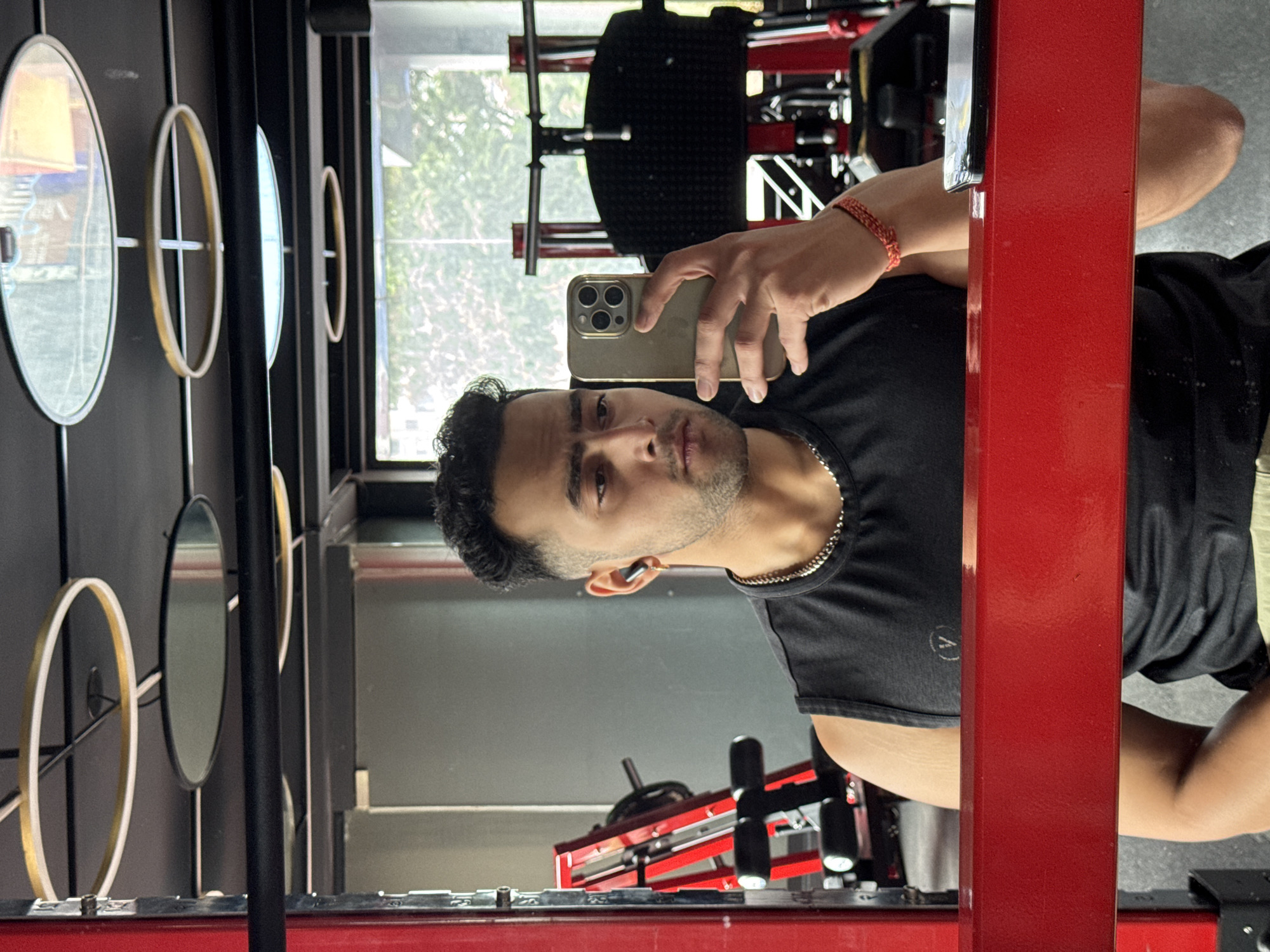 A young Indian man is taking a mirror selfie in a gym. He is wearing a black sleeveless t-shirt and has earbuds in his ears. He is holding a gold iPhone in his right hand, and his left hand is visible in the frame. A red metal bar is in the foreground, partially obscuring the man's lower body. The background shows gym equipment, including a red weight rack and other machines. The ceiling has decorative circular light fixtures. The image is taken from a medium shot, focusing on the man and his reflection. There are no clear indicators of the specific location within Kolkata, India.