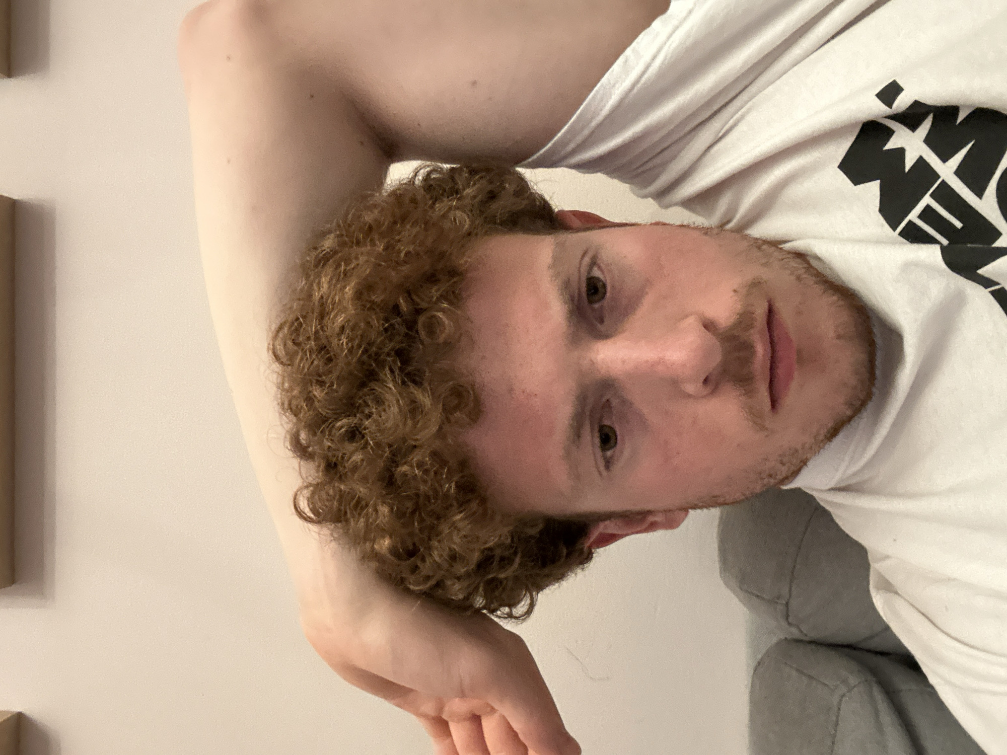The image shows a person with curly, reddish-brown hair, fair skin, and a light beard, lying down with their arm raised. They are wearing a white t-shirt with a black graphic on the right side of the chest. The background is a light neutral color, and a portion of a gray upholstered couch is visible on the lower right. There is also a light-colored wall with geometric panels in the upper left. The image appears to have been taken indoors with artificial lighting. There is no information in the image to suggest that it was taken in New York City, United States of America.
