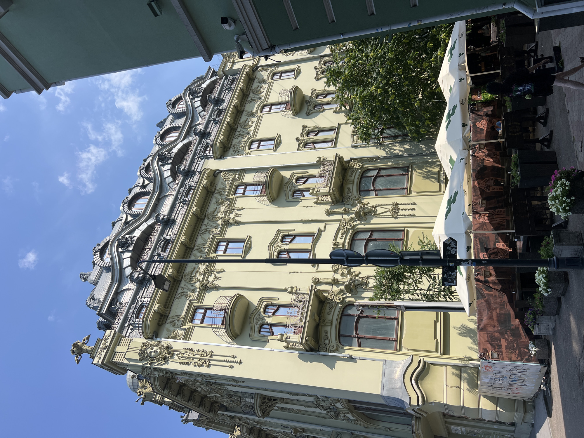 The image shows the exterior of a historic building in Odesa, Ukraine, with an Art Nouveau facade, located on a sunny day. The building is predominantly pale green with ornate decorative elements, including sculptures and intricate carvings. Several windows with dark frames are visible, some with small balconies. The roof is characterized by a distinctive mansard style with dormer windows. In front of the building, there's an outdoor seating area of a cafe or restaurant, with white umbrellas and tables. A street lamp stands prominently in the foreground. A person, a woman, is walking past the seating area on the sidewalk. Adjacent to the ornate building, a modern, plain green building is partially visible on the right side of the frame. The sky is blue with scattered clouds.