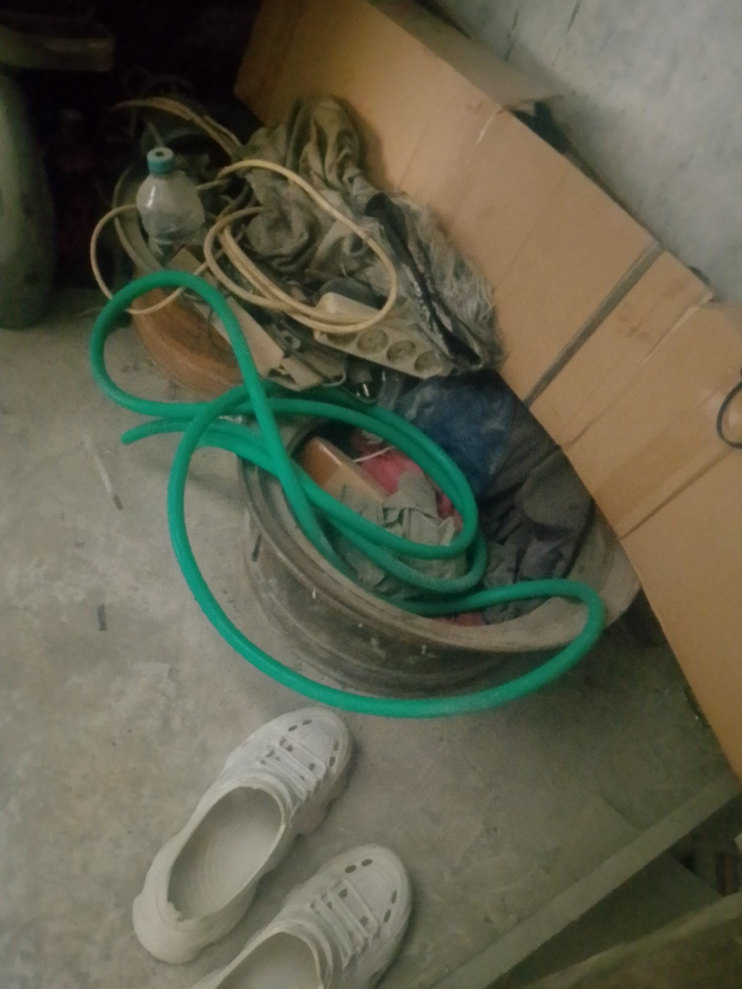 This image depicts a cluttered indoor space, possibly a storage area or a workshop, on a concrete floor. In the foreground, a pair of white sneakers are visible. To the left and center, a green hose is coiled within a metal rim or basin, surrounded by various objects including a clear plastic bottle with a blue cap, and coiled electrical cords. In the background, stacks of cardboard boxes are present, leaning against a wall. The lighting is dim, suggesting an indoor environment. There is no indication of the specific location in Banjarmasin, Indonesia, as the image lacks any identifiable landmarks or context. The scene appears to be static, with no people or animals present.
