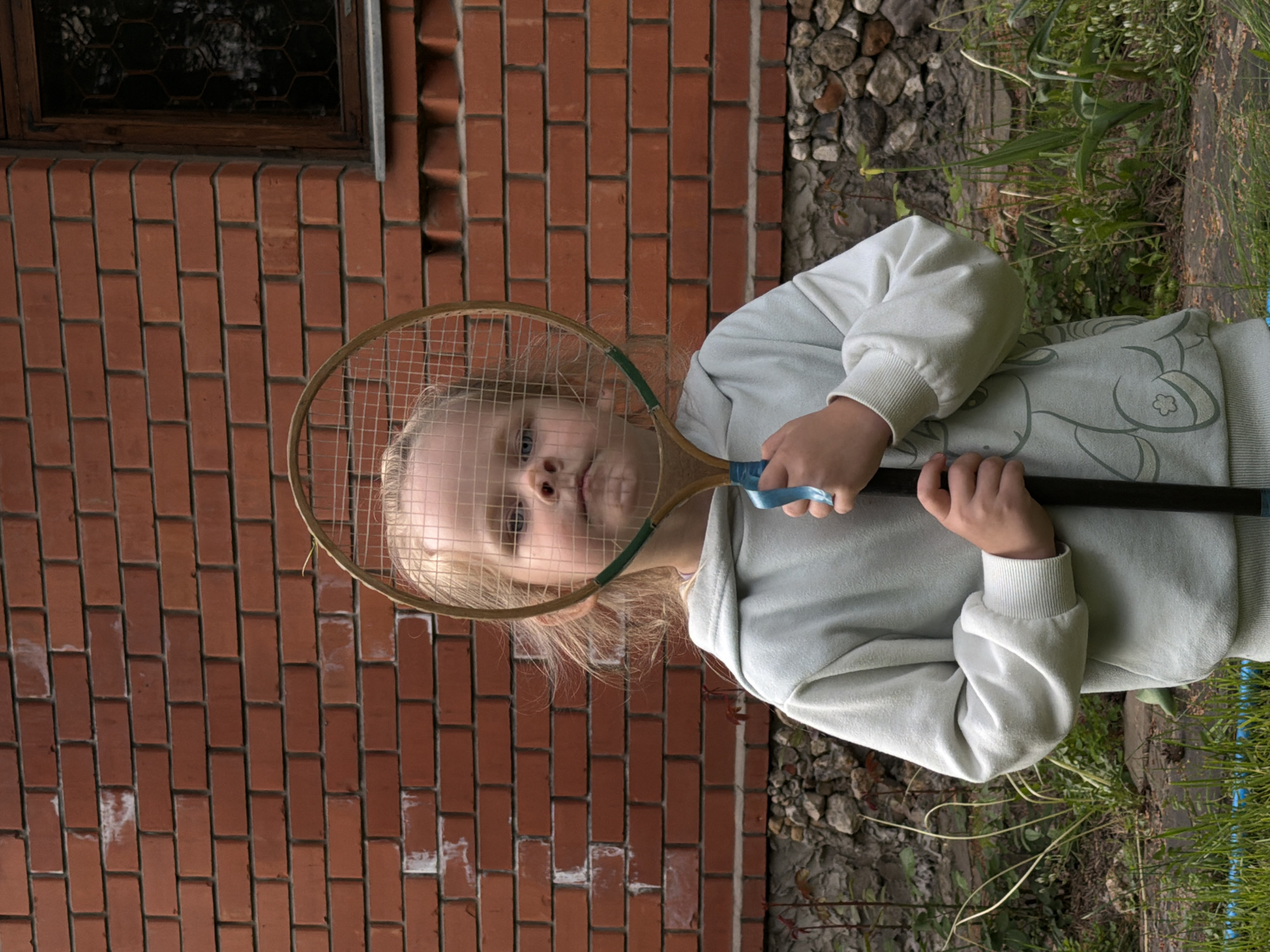 A young girl is outside, holding a badminton racket in front of her face. The girl has fair skin, light blonde hair, and is wearing a light-colored hooded sweatshirt. Behind her is a red brick wall with a window and an area of stones and greenery. The photograph was taken outdoors in Udelnaya, Russia.