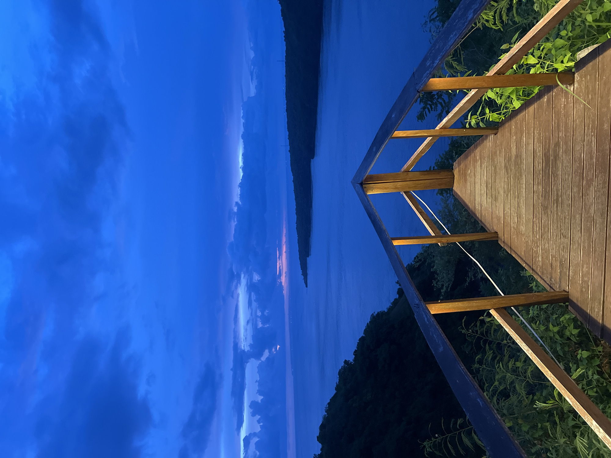 The image captures a serene scene at night in Nusa Penida, Indonesia. It is taken from a wooden deck overlooking a body of water, possibly a bay or inlet, with land visible on both sides. The sky is a deep blue, with hints of pink and purple suggesting dusk. Clouds are scattered across the sky. The wooden deck has a railing made of wood and green foliage growing around the edges. A white wire runs along the deck's railing. The overall impression is one of tranquility and natural beauty.
