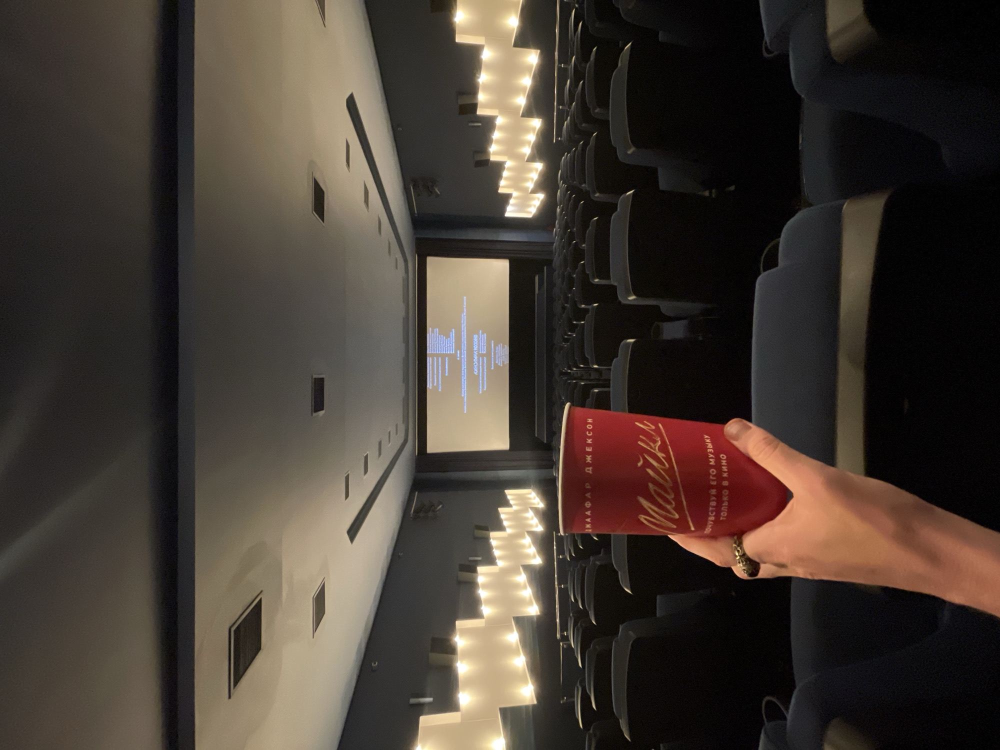 The image shows a person holding a red cup with text in Russian, in a dark movie theater. The cup has the text "МАЙКИ" in a script font, and below that, "ПОЧУВСТВУЙ ЕГО МУЗЫКУ ТОЛЬКО В КИНО". The screen at the front of the theater is illuminated, displaying credits. The seats are dark blue, and the walls have a decorative element with lights. This scene is likely in Saint Petersburg, Russia, as indicated by the Russian text on the cup.