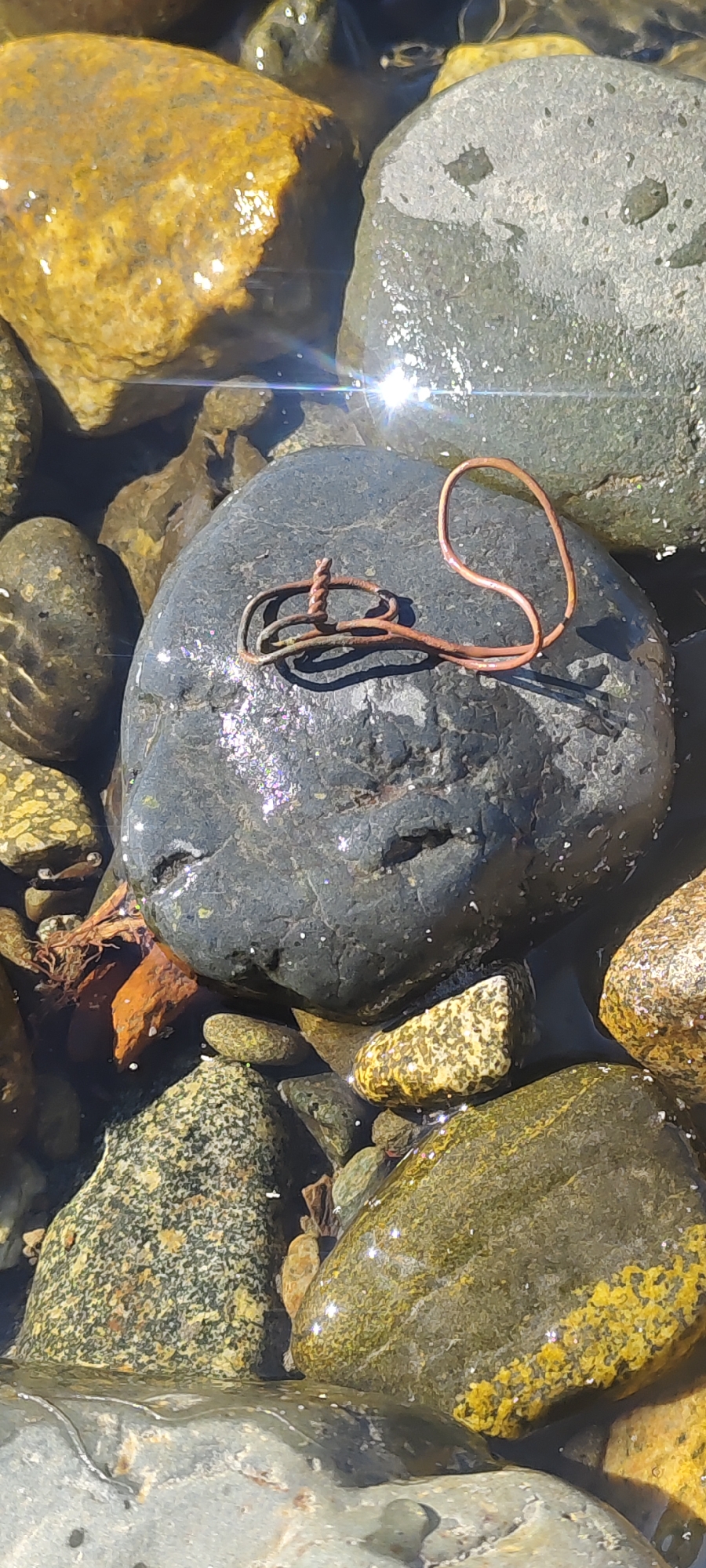 The image shows a close-up view of a dark-colored, round rock resting in shallow, clear water among other rocks and pebbles. A thin, rusty-colored metal object, possibly wire or a piece of jewelry, is placed on top of the dark rock. The metal object is bent into an irregular shape, with one end appearing twisted. Sunlight glints off the water and the surfaces of the rocks, creating bright reflections. The scene appears to be a natural setting, likely a riverbed or shoreline, with no discernible urban elements to suggest a specific location within Mezhdurechensk, Russia. There are no people or animals visible in the image, and no actions or interactions are depicted. The focus is solely on the rock and the metallic object.