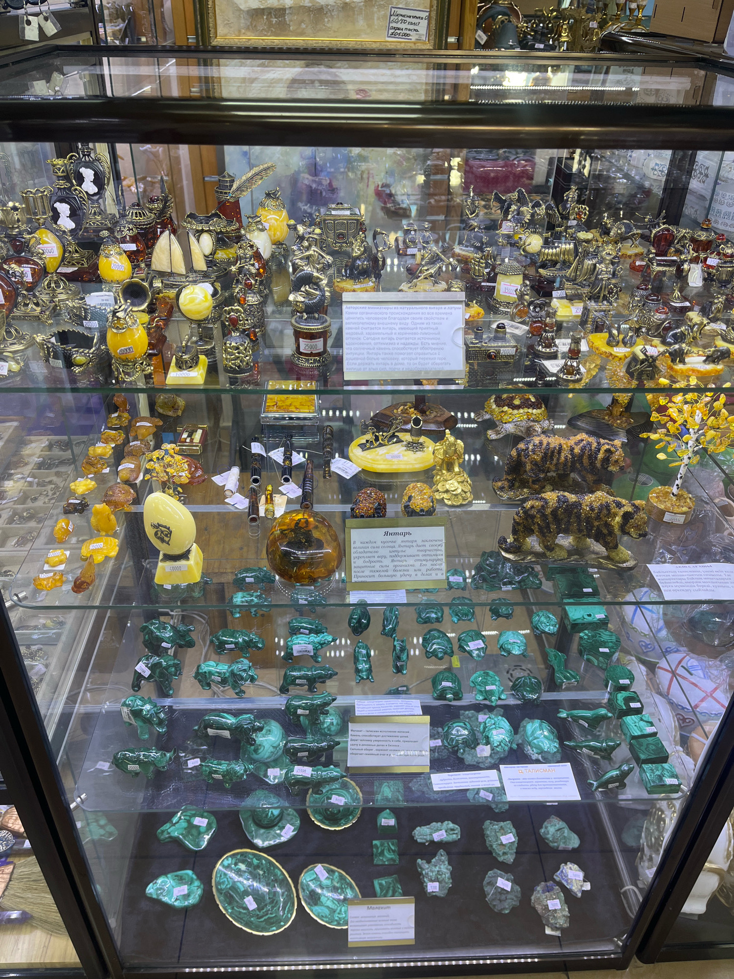 The image displays a well-lit glass display case filled with various decorative items, likely in a store. The items are primarily made of polished stones, metals, and amber, and include figurines of animals such as tigers, bears, horses, and seals, as well as decorative eggs, bowls, and abstract sculptures. Some items have small price tags attached. The upper shelves contain more intricate, metallic pieces, including what appear to be decorative vessels and small figurines. The lighting within the case is bright, casting reflections on the glass and the polished surfaces of the objects. The background visible through the glass is blurred, suggesting a commercial environment. There is no direct indication in the image that this location is in Astana, Kazakhstan, nor are there any people present.