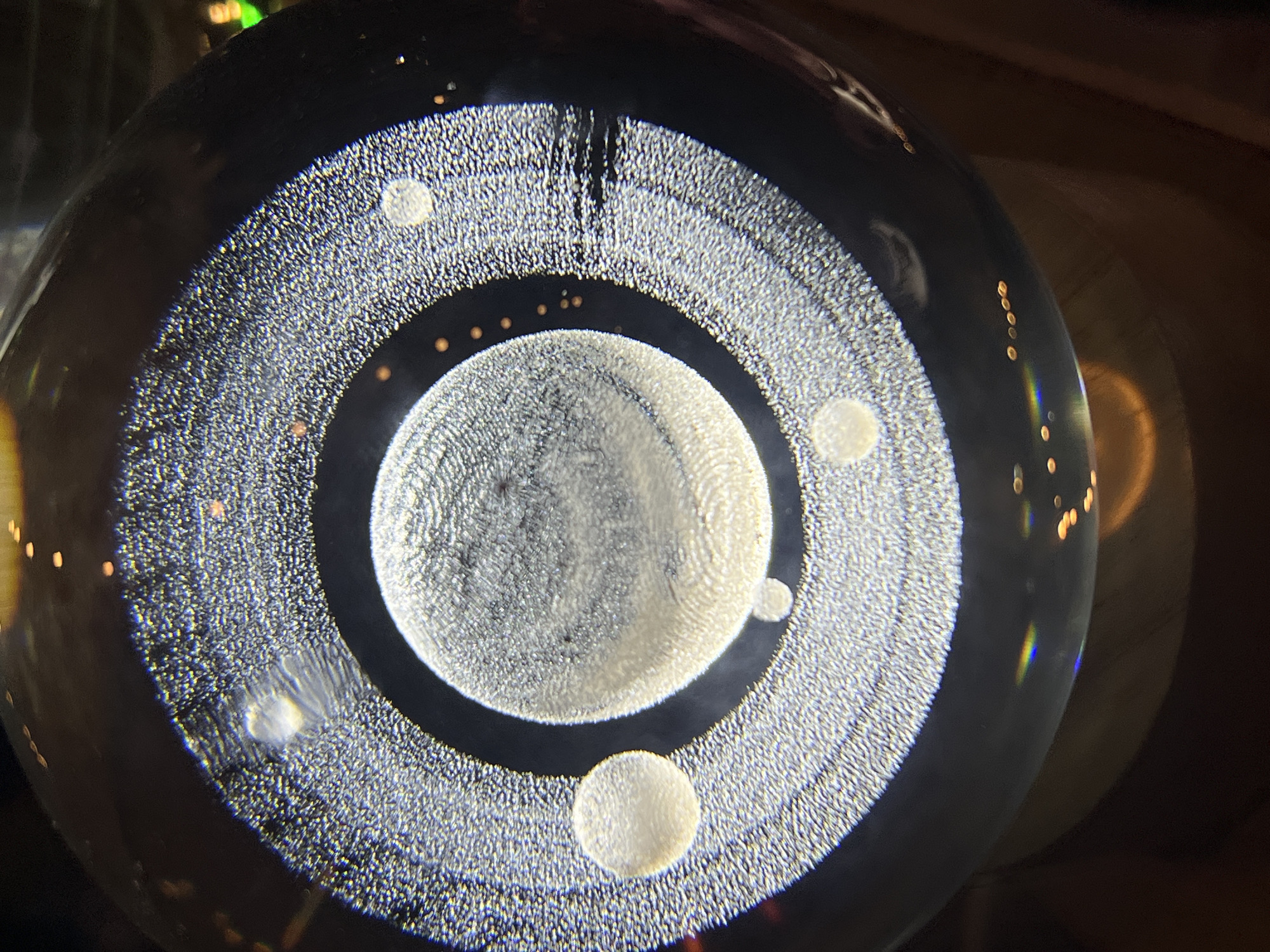 The image depicts a glass orb containing a pattern resembling a celestial body or a stylized planetary system. It features a central sphere with a grainy texture, surrounded by concentric rings of similar texture, and several smaller circular "satellite" shapes interspersed within the rings. The orb appears to be reflecting light, causing some areas to appear brighter. The background is dark and blurred, possibly indicating a low-light setting. There is no indication in the image to suggest the location is Unknown, Unknown.