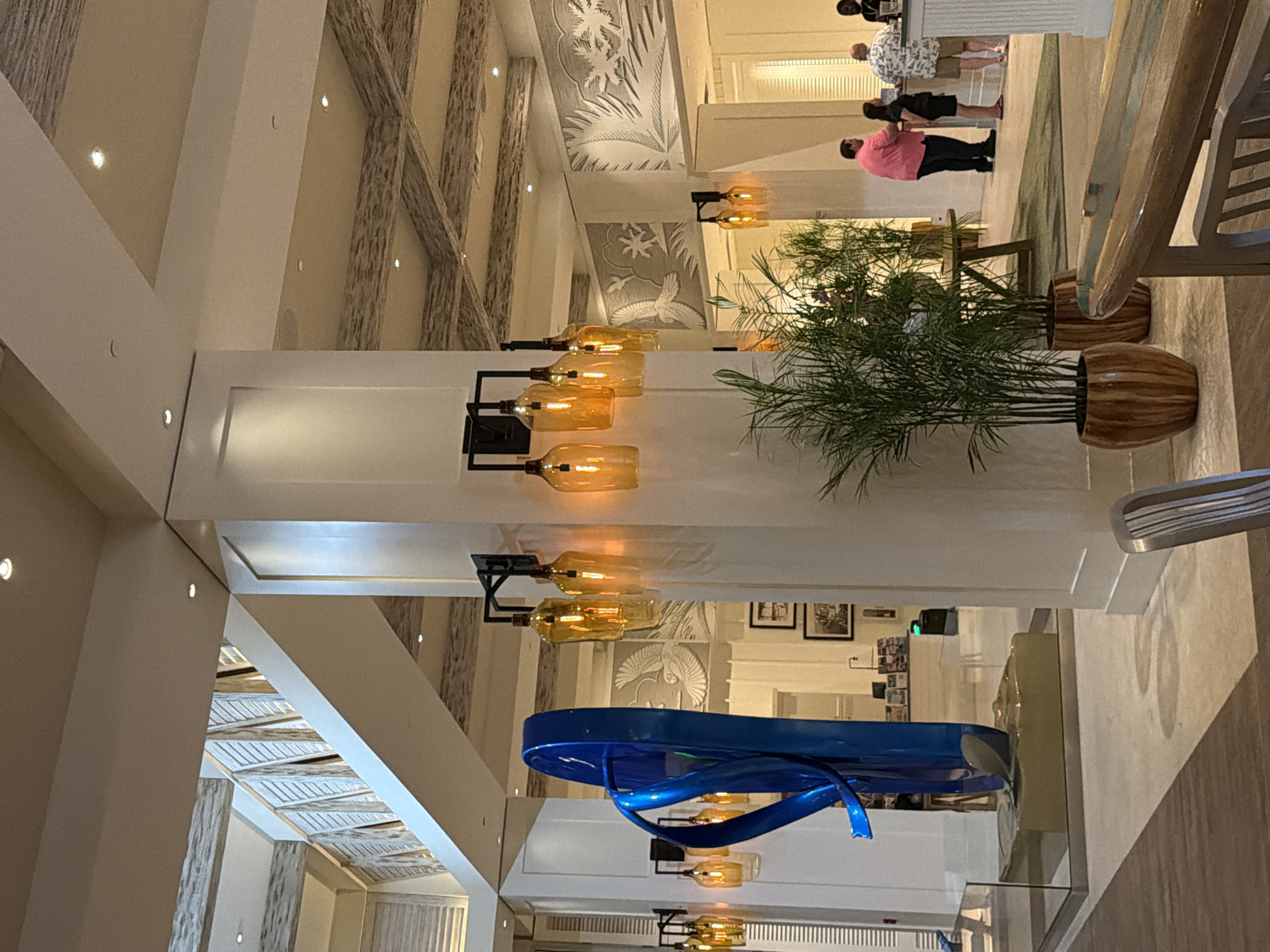 The image depicts the interior of a hotel lobby in Hollywood, United States of America. The scene is characterized by a large, blue, abstract sculpture of a flip-flop positioned near the entrance. The lobby features a decorative ceiling with exposed beams and recessed lighting. Wall sconces with amber-colored glass globes are mounted on a large white pillar, casting a warm glow. Several potted plants, including tall, slender palm-like plants in wooden planters, are visible. In the background, a reception desk is partially visible, with several individuals interacting at the desk, including a person in a pink shirt, a person in a black dress, and a person in a patterned shirt and shorts. The overall ambiance suggests a modern, tropical-themed hotel.