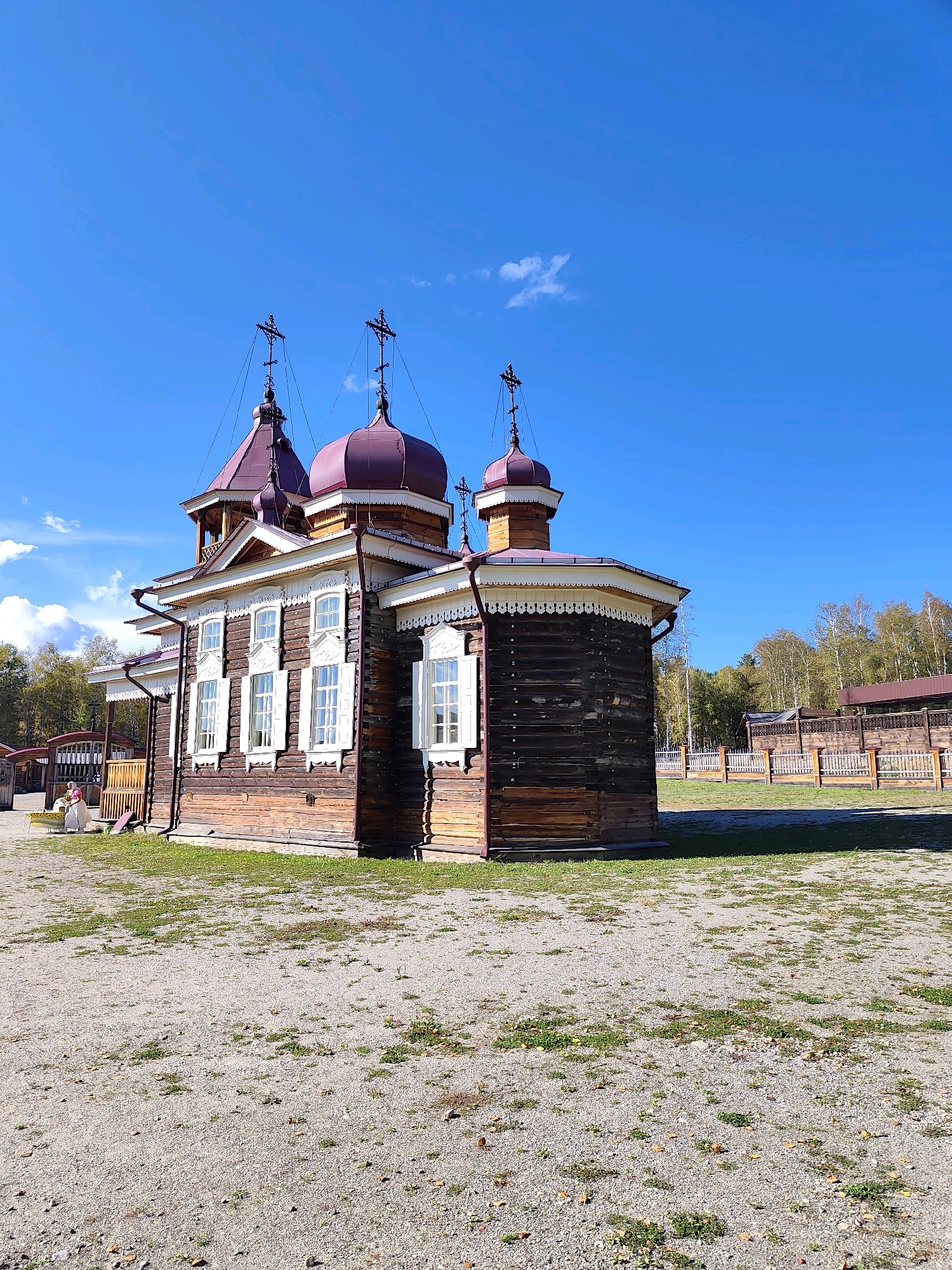 A wooden Orthodox church with a maroon dome and cross stands in the foreground. The church is made of dark logs and features white-framed windows with open shutters and decorative white trim along the roofline. To the left of the church, a woman in a pink headscarf is seated on a low bench next to a yellow object, possibly a basket. Beyond the church, a wooden fence encloses an area with a few trees, some with yellowing leaves, suggesting it is autumn. The ground in front of the church is covered with gravel and scattered green vegetation. The sky is a clear, bright blue with a few wispy white clouds. The scene appears to be located in a rural or historical settlement setting, consistent with the ethnographic museum of Taltsy, Irkutsk, Russia.
