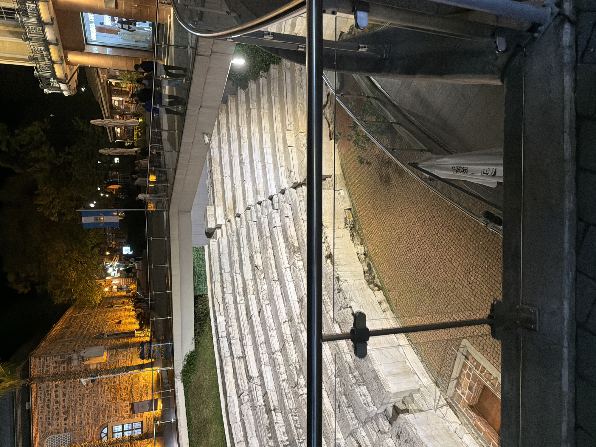 The image captures an illuminated urban scene at night in Plovdiv, Bulgaria, showcasing a blend of historical architecture and modern public spaces. The perspective is from an elevated viewpoint, looking down onto a multi-level area.
In the foreground, a modern glass and metal railing system, likely part of a bridge or overlook, divides the scene. Beyond this railing, a prominent feature is a large, tiered stone structure, resembling an ancient amphitheater or stadium seating, extending across the midground. This structure is brightly lit by artificial lights, highlighting its aged, rugged appearance. Adjacent to it on the right, an area of textured cobblestone pavement is visible.
To the left side of the image, an older building with distinctive patterned brickwork and arched windows stands out, illuminated by warm lights. Trees and streetlights are scattered in this area, along with a flag featuring horizontal blue and white stripes. Further into the background, several outdoor cafes or restaurants are visible, with people sitting at tables under umbrellas.
On the right side of the image, another building features a large display window with partially visible text, "GONDEL GROF." More people are seen in this area, some dining outdoors or walking. A white umbrella, possibly belonging to one of the establishments, displays faint text including "CRISTAL" and a QR code.
The overall atmosphere is lively and inviting, characteristic of an evening in a bustling historical city. The dark sky contrasts with the numerous artificial light sources, which cast a warm glow over the various architectural elements and social activities taking place.