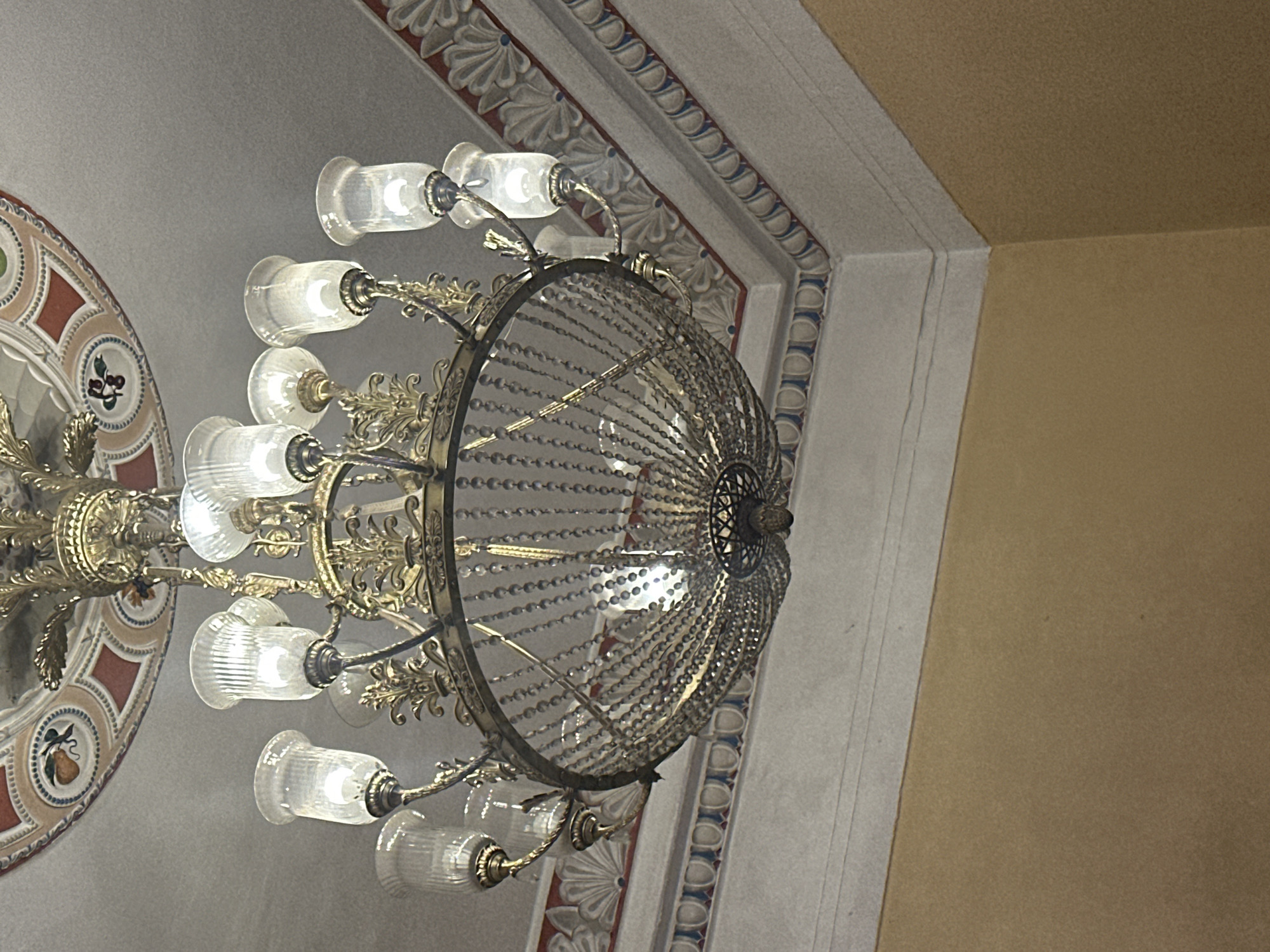 An ornate, multi-tiered chandelier made of brass and crystal hangs from a decorated ceiling in what appears to be a public building. The chandelier has multiple glass sconces, each containing a light bulb. The ceiling is painted with intricate patterns and borders, featuring floral motifs and geometric designs in shades of red, brown, and blue. The walls below the ceiling are a muted yellow. The image is taken from a low angle, looking up at the chandelier and ceiling. There are no people visible in the image. The scene is static and well-lit, suggesting the chandelier is functional. The architectural style of the ceiling and the chandelier's design imply an older, possibly historic, building. Without further information, it is not possible to determine the exact location in Minsk, Belarus, though the style could be indicative of a palace, museum, or grand theater.