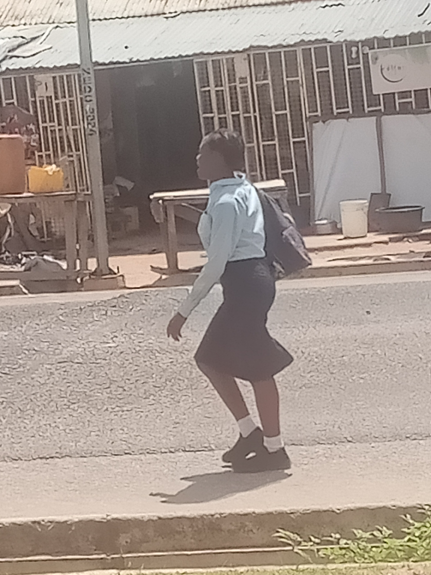 A person wearing a light blue long-sleeved shirt and a dark skirt walks on a paved surface next to a road. The person carries a dark backpack over their right shoulder. The person is on the left side of the image, facing right. In the background, there is a building with a corrugated metal roof and a fence. A sign with a logo is visible on the building. There are various objects on the ground and near the building, including buckets and what appears to be a shovel. A pole with writing on it is located to the left of the person. The ground is covered in gravel. The scene appears to be in an urban or semi-urban environment, possibly in Jalingo, Nigeria.