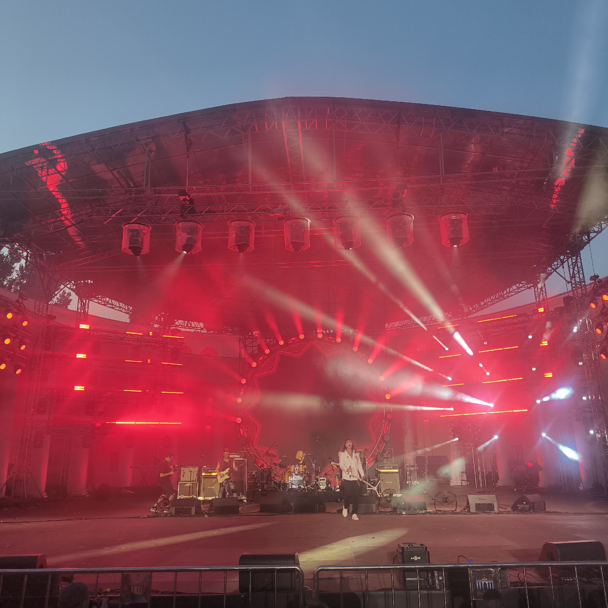 An outdoor stage is illuminated by red lights and haze. Several people are visible on the stage, including a lead singer in a white shirt and dark pants who is walking across the stage. To the left of the singer, a person is playing a guitar. Behind them, a drummer is seated at a drum set. Various amplifiers, speakers, and lighting equipment are positioned on the stage. The stage is covered by a large metal structure that supports a canopy. In the background, there are trees and a building. The sky above is clear and a light blue. The image appears to be taken at dusk.