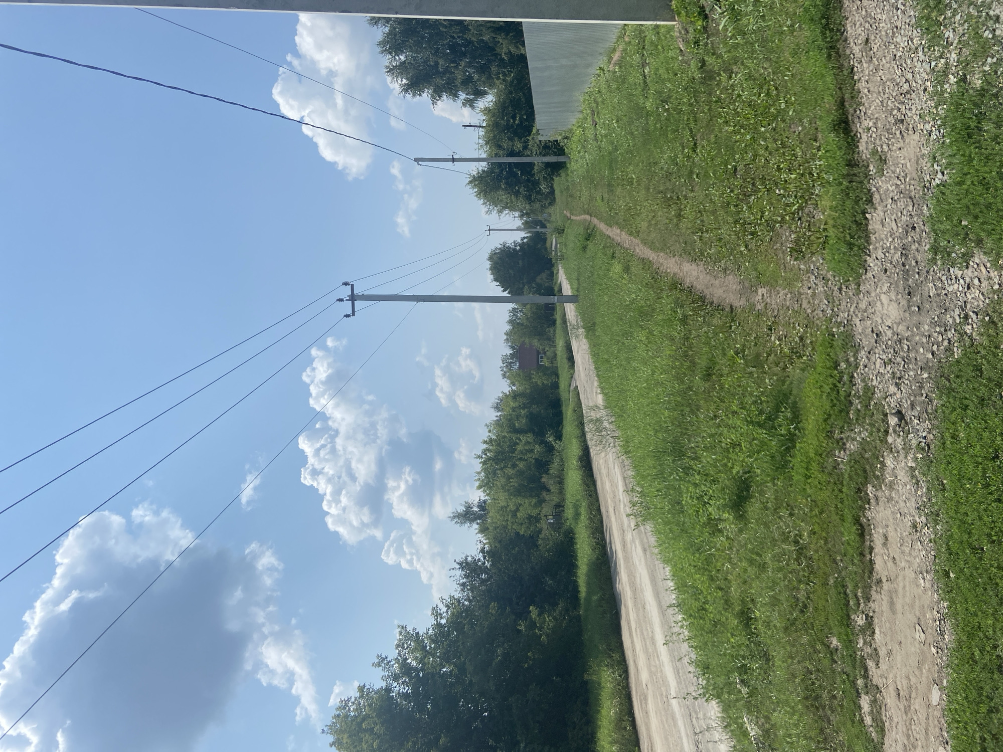 The image depicts a rural or suburban scene on a clear, sunny day. A dirt road curves to the left, bordered by lush green grass and trees on either side. Overhead, power lines stretch across the bright blue sky, supported by utility poles. In the distance, a small house is visible amongst the trees. A narrow dirt path runs parallel to the road, leading into the distance. A corrugated metal fence lines the right side of the scene. The overall atmosphere is tranquil and natural, suggesting a quiet, undeveloped area.