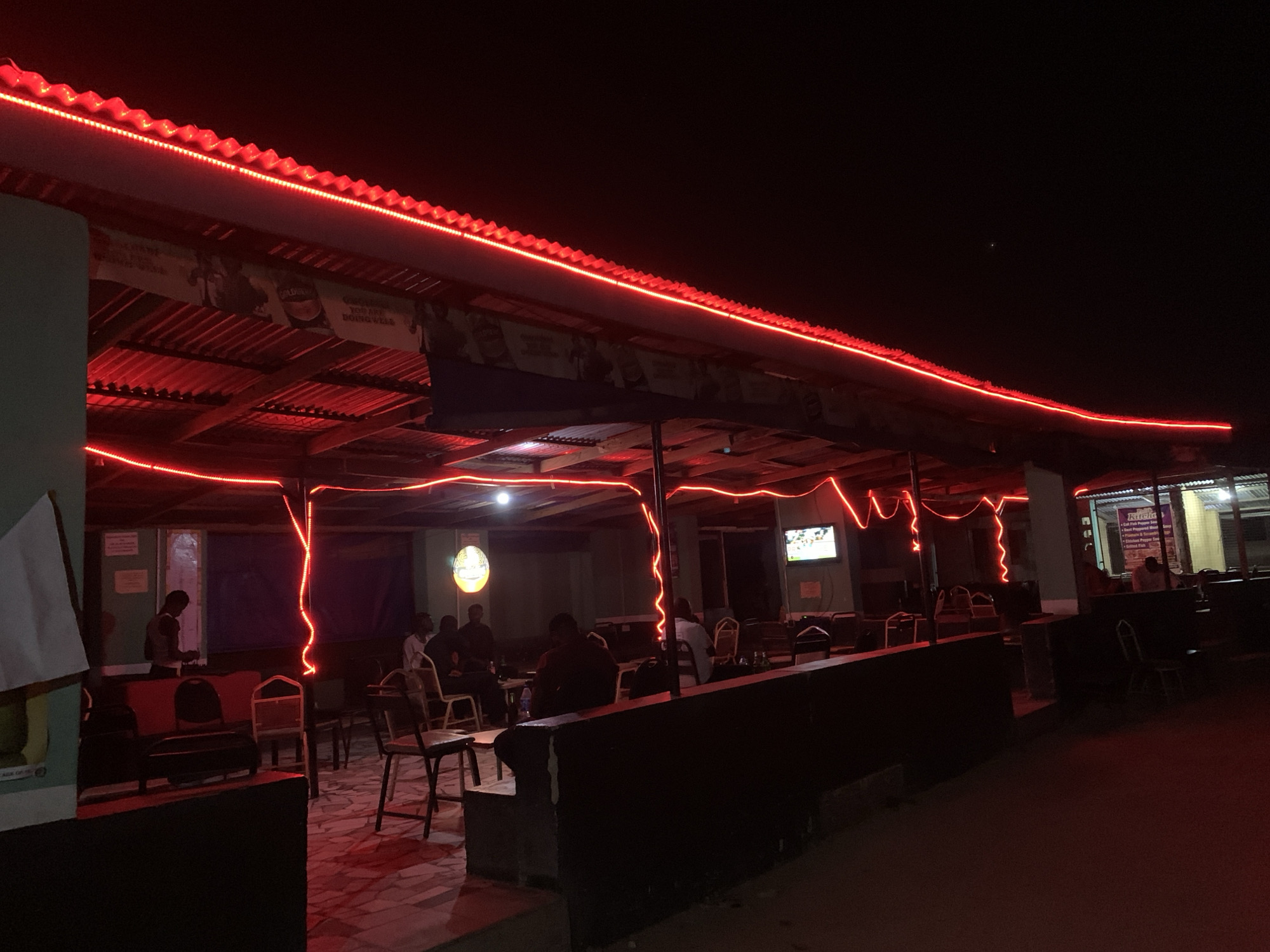 The image shows an outdoor bar or restaurant in New Stadium, Nigeria, at night. Red LED strip lights outline the edge of the corrugated metal roof. Several people are sitting at tables inside the covered space. A television is visible, mounted on a wall inside. The bar appears to be dimly lit. There is a partially obscured sign at the entrance. The ground outside is unpaved.
