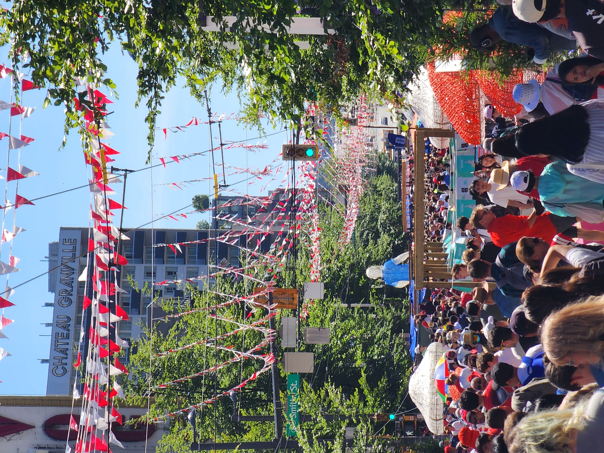 In an urban outdoor scene, a bustling street fair is in progress under a bright, clear sky. Red and white bunting stretches across the street, adorning trees and lampposts. A multistory building with "CHATEAU GRANVILLE" signage is visible in the background. Dense crowds of people, dressed in casual summer attire, fill the street, creating a vibrant and energetic atmosphere. In the foreground, a large, blue, sculptural figure stands above a wooden structure, partially obscured by the crowd. Traffic lights show a green light. A street sign indicates "Smith St.". Numerous vendor stalls and tents are set up along the street, suggesting a lively marketplace or community event. The scene is filled with a sense of festivity and community gathering.