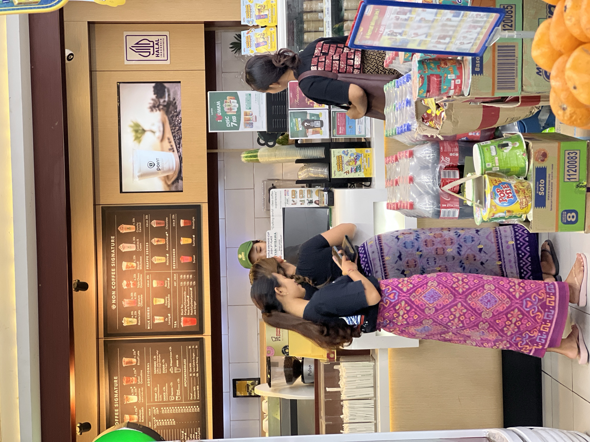 The image captures a bustling scene inside a cafe or convenience store located in Celuk, Indonesia. Three individuals are visible: one female customer stands near the counter, examining merchandise, and two others are behind the counter, interacting with each other. One of the two individuals behind the counter is wearing a green hat. The cafe features a prominent menu board displaying coffee and non-coffee options, along with their prices. Shelves are stocked with various items, including bottled drinks, instant noodle cups, and other packaged goods. The store displays a "HALAL Indonesia" logo. Bright lighting and a variety of products create a busy retail environment.