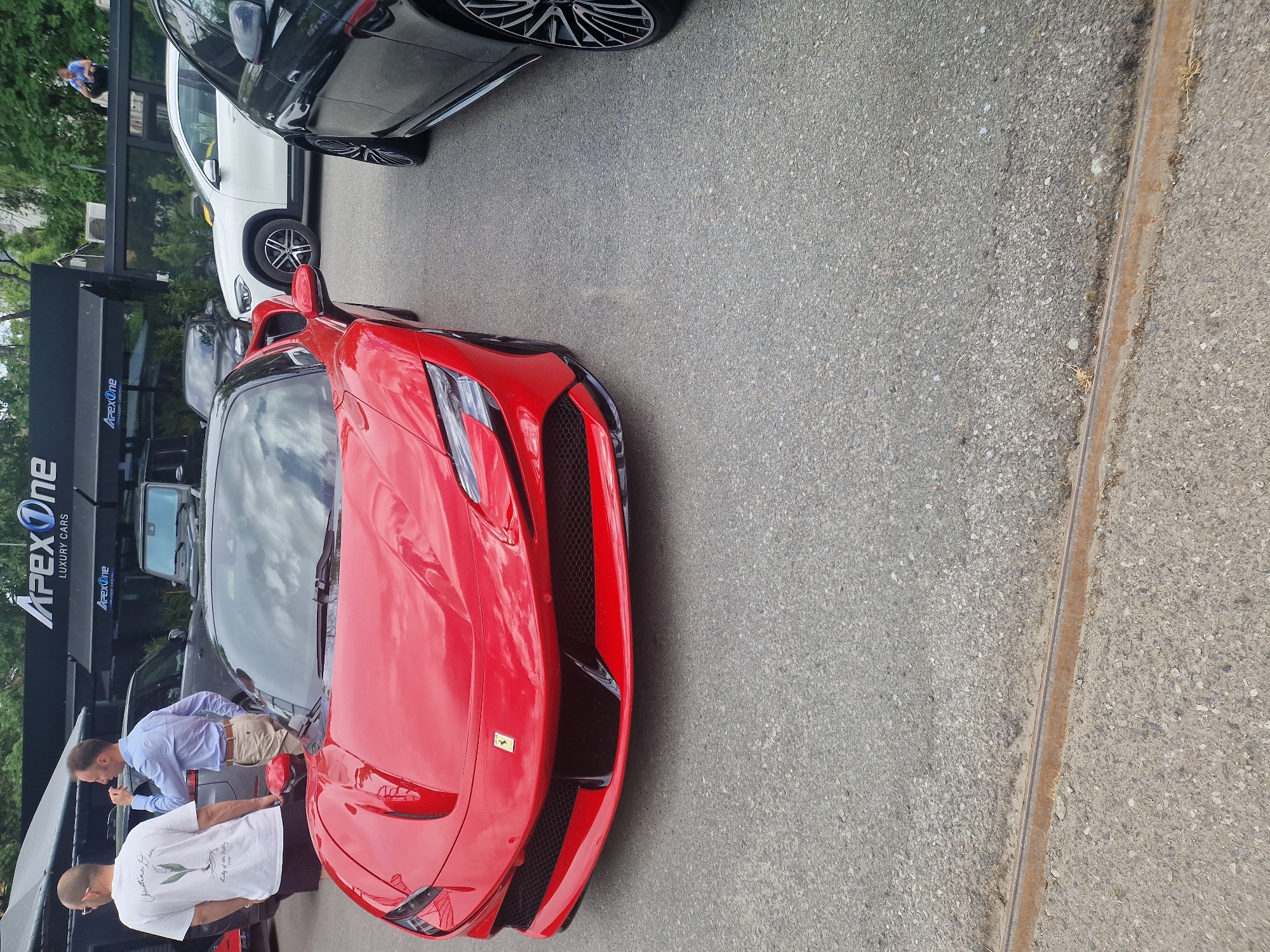The image depicts a red Ferrari parked on asphalt, likely at a luxury car dealership. Two men are standing near the front of the vehicle, seemingly examining it. One man is wearing a white t-shirt and black pants, while the other is wearing a light blue button-down shirt and khaki pants. Other luxury cars are visible in the background, parked near a building with the sign "Apex One Luxury Cars." The location appears to be an urban environment. This image was taken in Sofia, Bulgaria.