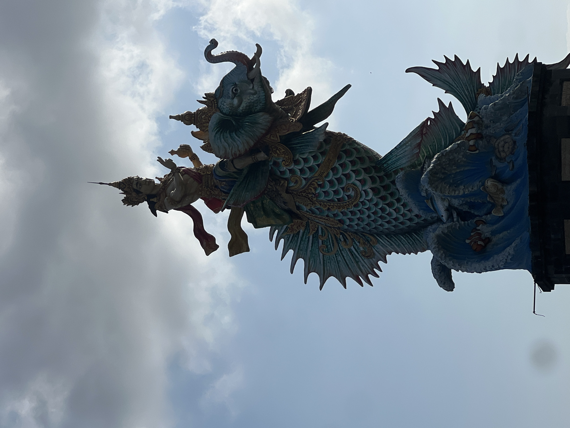 A statue of a mythical creature is silhouetted against a cloudy sky in Mengwi, Indonesia. The creature has the body of a fish, with scales and fins, and a head resembling an elephant. A figure, adorned in a crown and flowing robes, sits atop the elephant's head, holding an object in its left hand and gesturing with its right. The statue is supported by a base that appears to depict crashing waves with fish and sea creatures. The sky is partly cloudy, with lighter clouds scattered against a pale blue background.