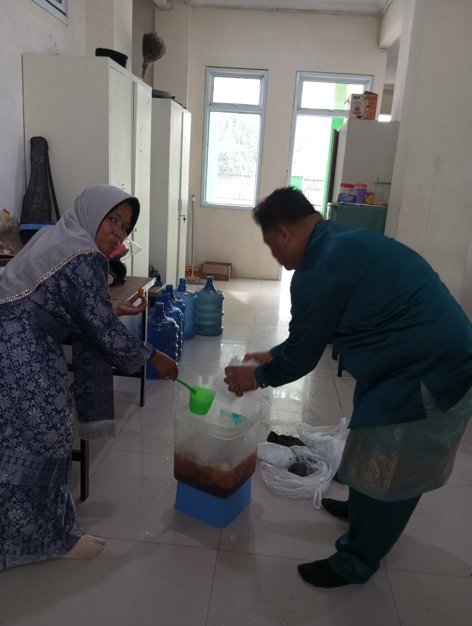 In a room with tiled floors and pale walls, two individuals are engaged in an activity involving a large plastic container filled with liquid and ice. A woman wearing a grey hijab and a patterned blue dress is standing on the left, holding a piece of food in her right hand and a green ladle in her left, which is positioned over the container. She is looking towards the camera. On the right, a man wearing a teal colored shirt and pants is bending over the container, his hands are near the opening. Behind the woman, several large blue water jugs are lined up against a white cabinet. To the right of the man, a clear plastic bag with dark objects inside is on the floor. The room has a window in the background with natural light filtering through. Based on the attire of the individuals and the general appearance of the room, this scene is likely within Pematang Reba, Indonesia.