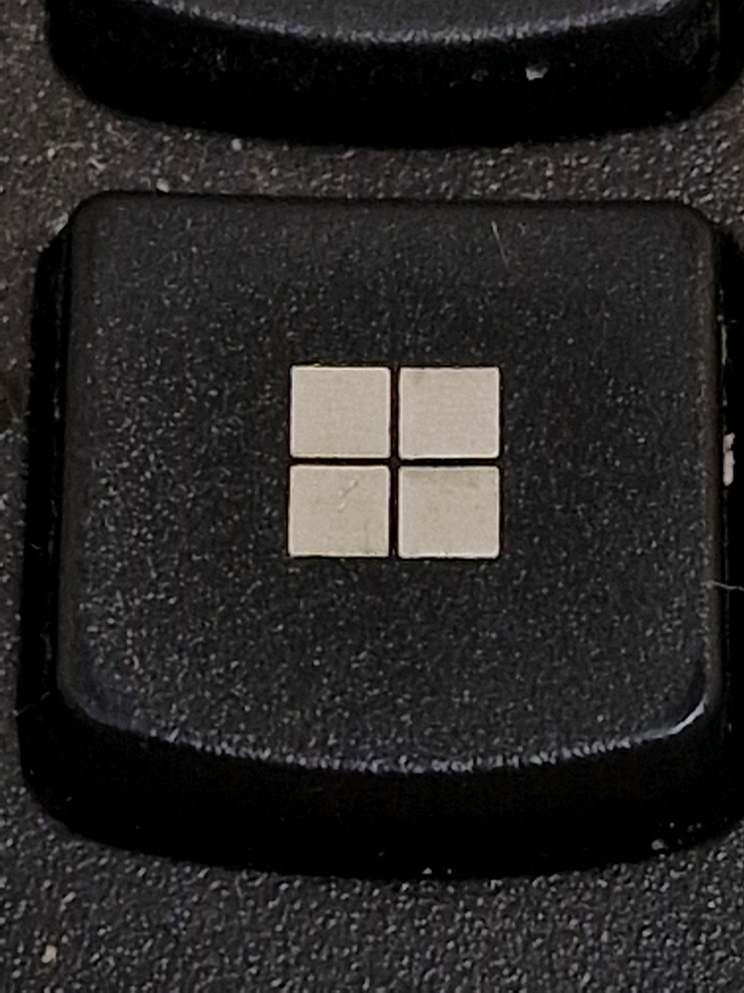 The image shows a close-up, top-down view of a black keyboard key with the Windows logo. The logo is comprised of four small, square, silver segments arranged in a 2x2 grid. The key is textured and appears worn. The background is out of focus and shows other parts of the keyboard. There is no indication of the city of Travnik, Bosnia and Herzegovina.