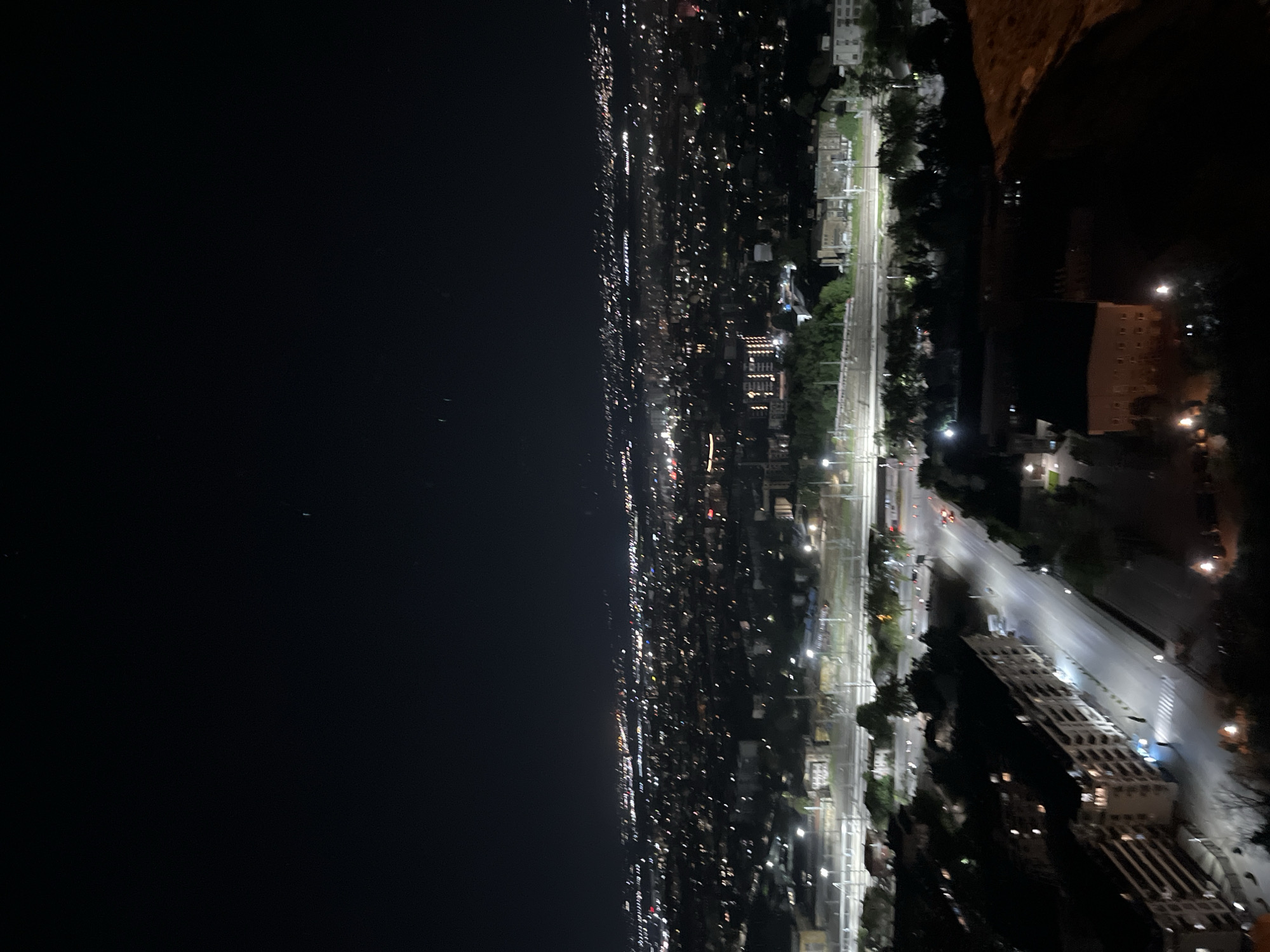 The image depicts a nighttime view of Plovdiv, Bulgaria, from a high vantage point, likely a hill or elevated structure. The city lights stretch across the horizon, contrasting with the dark sky on the left. A railway line runs parallel to a road with moving traffic, as indicated by the streaks of light. Buildings and trees are silhouetted against the illuminated streets and city lights. The foreground shows elements of the observation point, suggesting it is possibly a part of the Ancient Theatre of Philippopolis in Plovdiv.