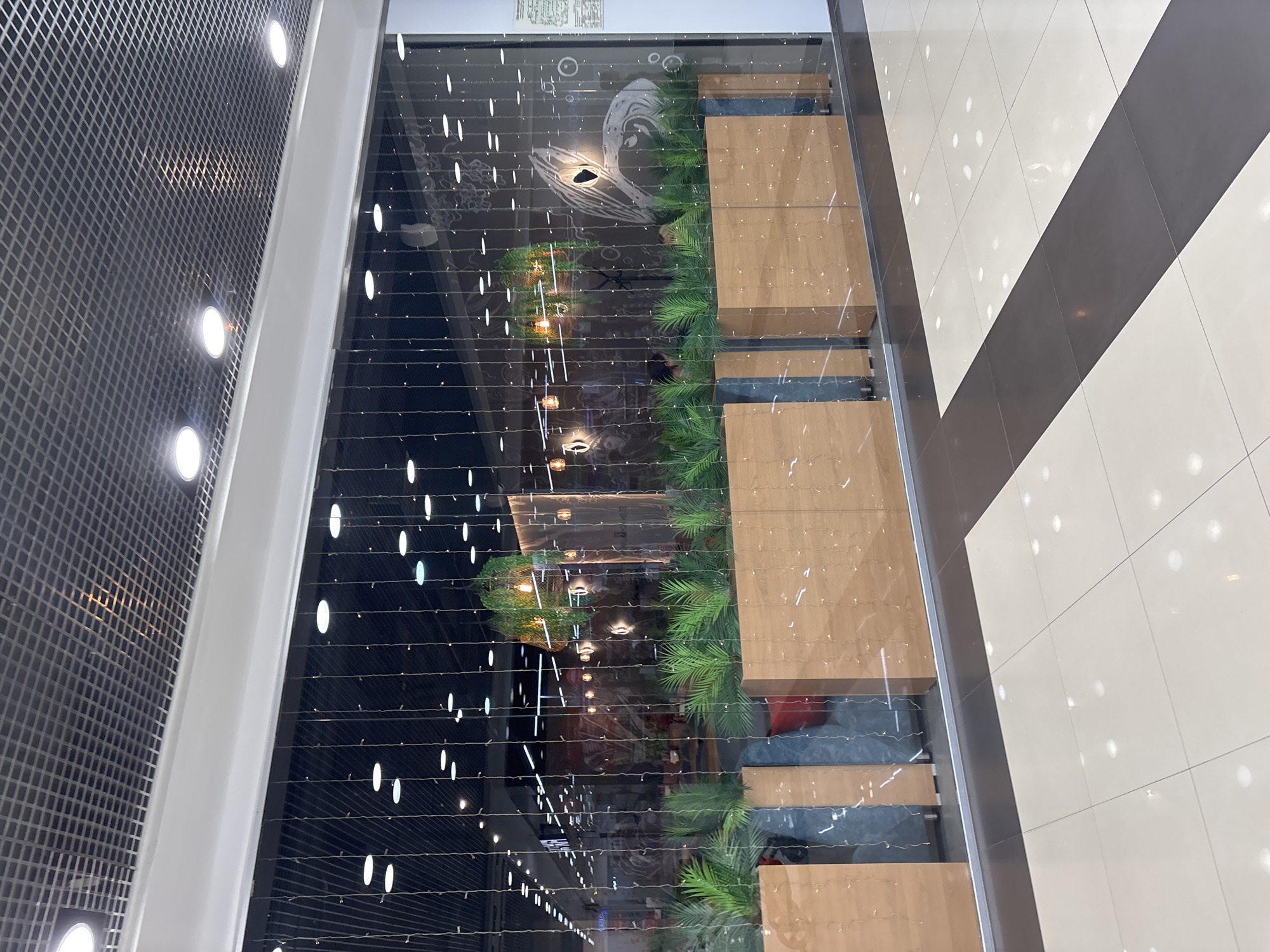 The image shows the interior of a shopping mall in Naberezhnye Chelny, Russia. The scene is dominated by the facade of a restaurant or cafe, visible through a clear glass wall draped with numerous vertical strings of small, warm-toned lights. Inside the establishment, plants are displayed in wooden planters, and decorative elements like hanging greenery and pendant lights are visible. The flooring in the mall is a patterned tile design with alternating light and dark grey squares. The ceiling features a grid of acoustic tiles with recessed circular lights. The overall atmosphere suggests a modern commercial space.