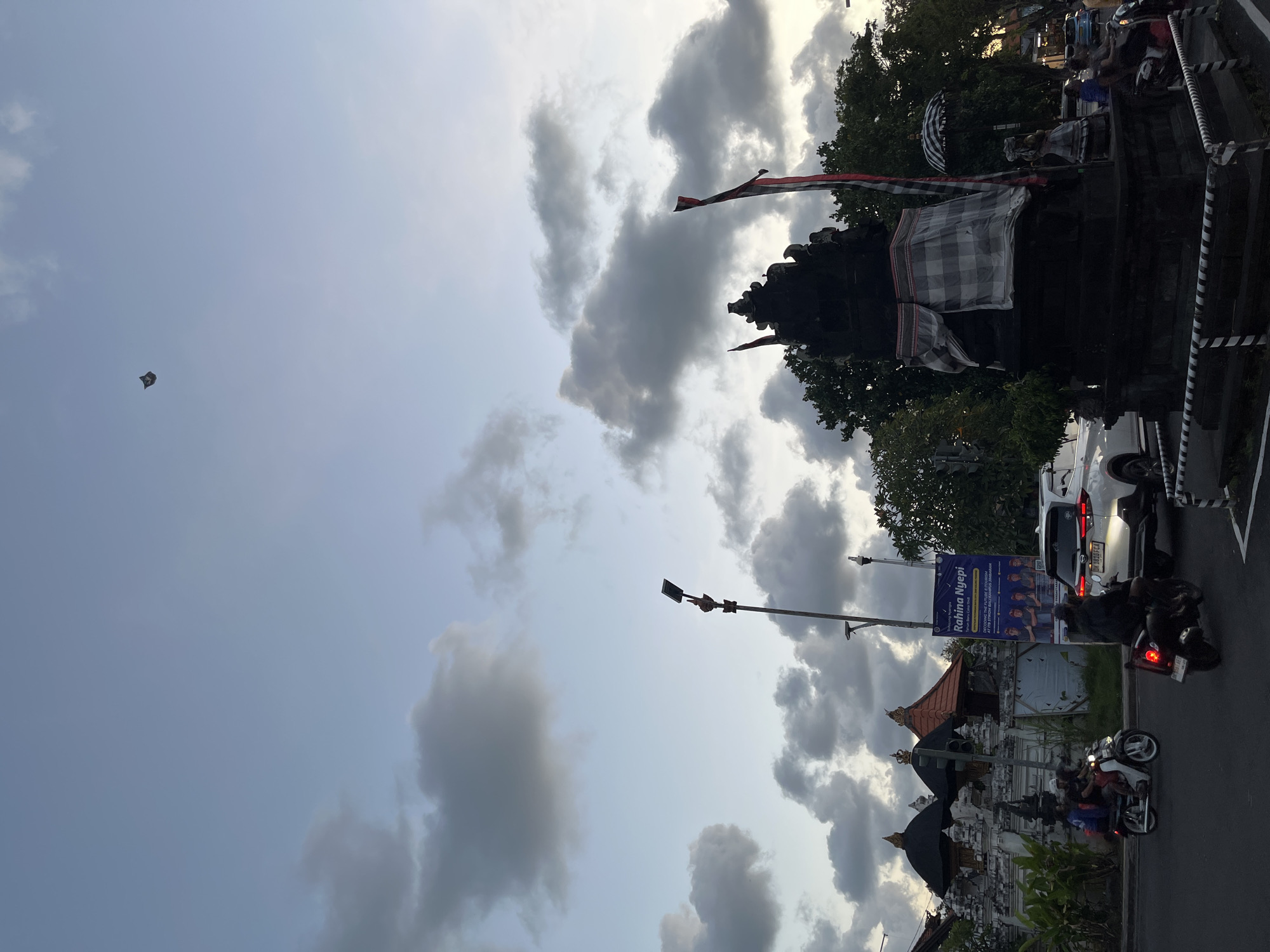 The photograph captures a streetscape in Kuta Selatan, Indonesia, presenting a mixture of local culture and daily life under a partly cloudy sky. A black kite is visible high up in the sky on the upper-left side of the frame. The lower portion of the image contains a portion of a stone structure, possibly a temple or shrine, draped with traditional Balinese cloth. An advertisement banner and street lights are situated nearby. A traffic scene is evident with motorcycles, cars, and people. The image is taken from a ground-level perspective, displaying the environment and surrounding details in a straightforward manner.
