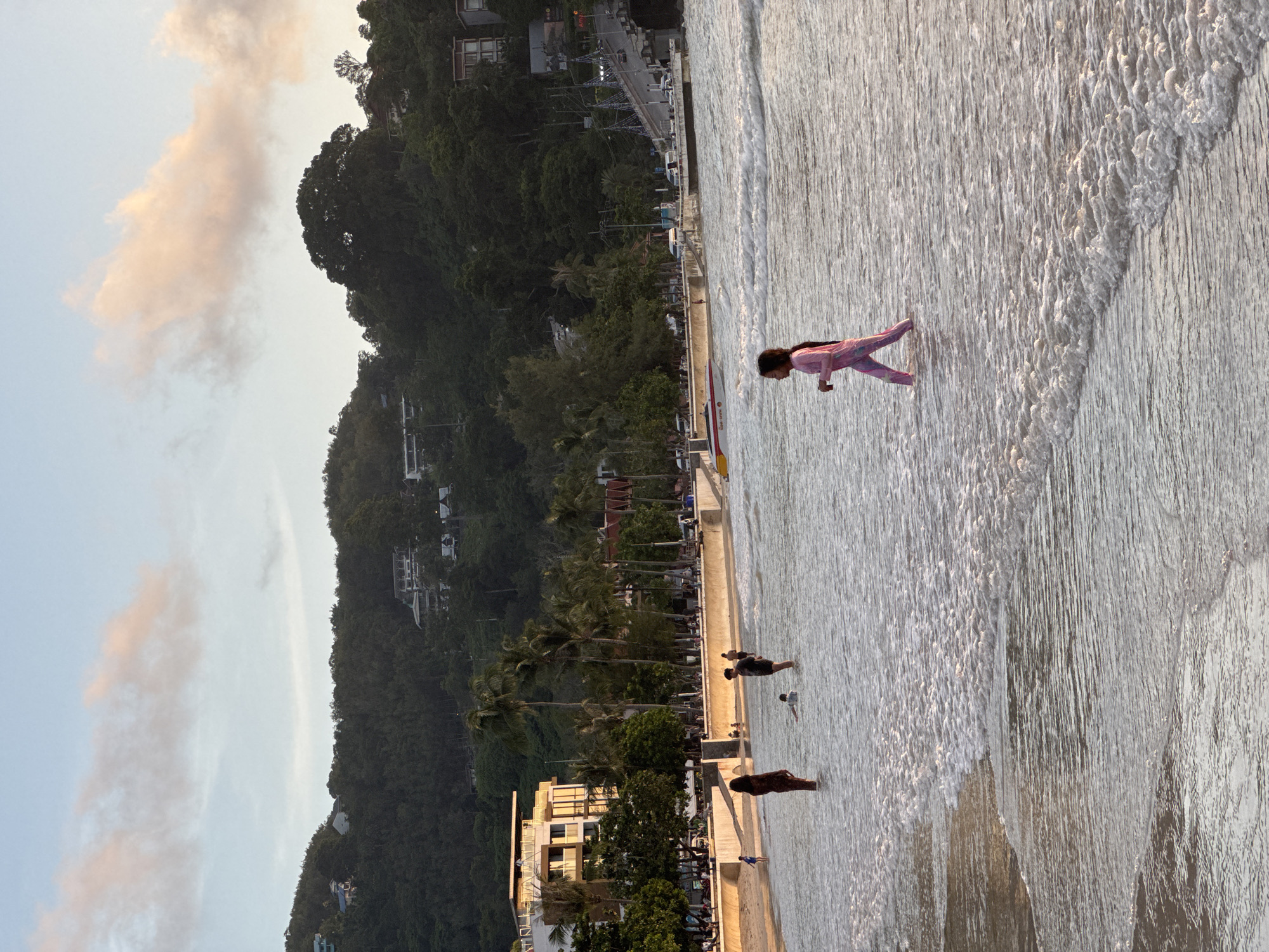 A wide shot captures the beach and ocean at Patong, Thailand, during the golden hour. The sky is a soft blue with hints of pink and orange clouds. In the foreground, the shallow ocean water is clear and wavy, with a young girl in a pink swimsuit walking to the right. Further back, three people stand in the water: a woman in a long, patterned dress, a man in dark shorts, and a child lying down in the water. The beach is visible behind them, lined with palm trees and buildings, including a modern hotel. In the distance, a lush green hill rises, dotted with houses and trees, and a bridge with traffic crosses a body of water. The overall scene depicts a serene evening at a popular tourist destination.