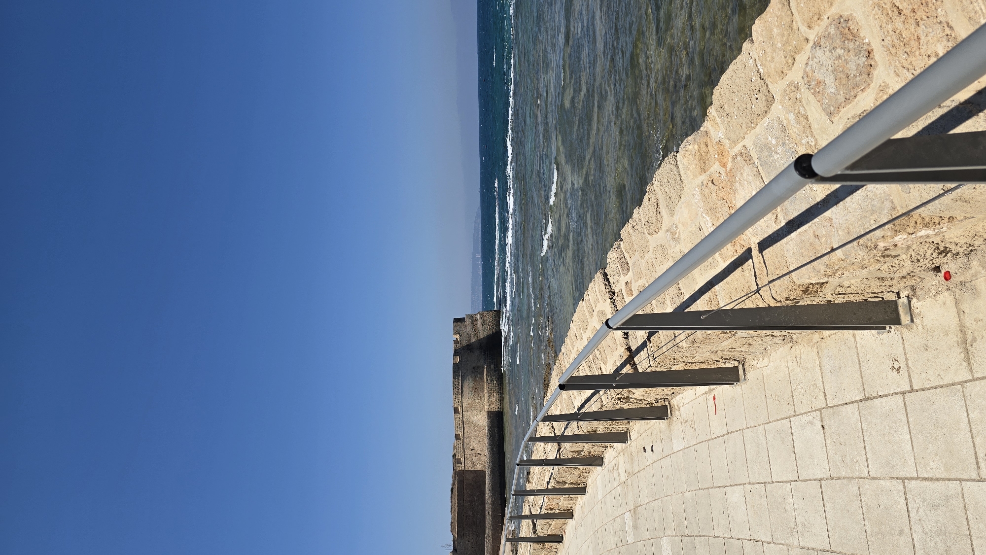 The image shows a view of the sea wall in the city of Acre, Israel. A paved walkway runs along the top of a stone wall, which has a metal railing. The sea is to the right, with waves breaking against the shore. In the background, a large stone fortress is visible. The sky is clear and blue.