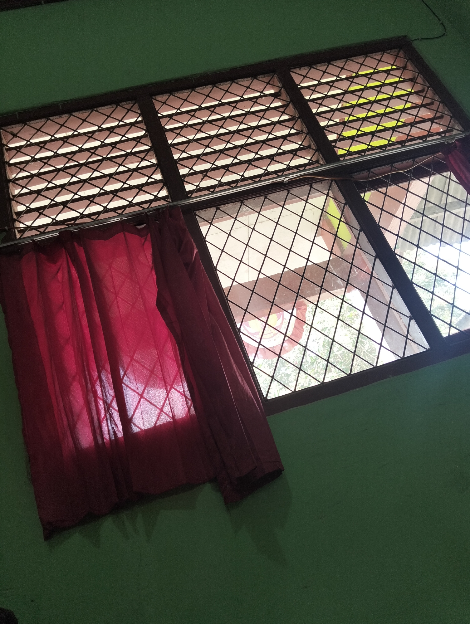 The image depicts a close-up of a window with a diamond-patterned security grate, partially covered by a deep red curtain. The window is set into a green-painted wall. Sunlight filters through the grate and the curtain, casting shadows. The scene appears to be indoors, with a view of the outdoors visible through the window. The location is identified as Banjar Agung, Indonesia. There are no people or specific actions visible in the image.