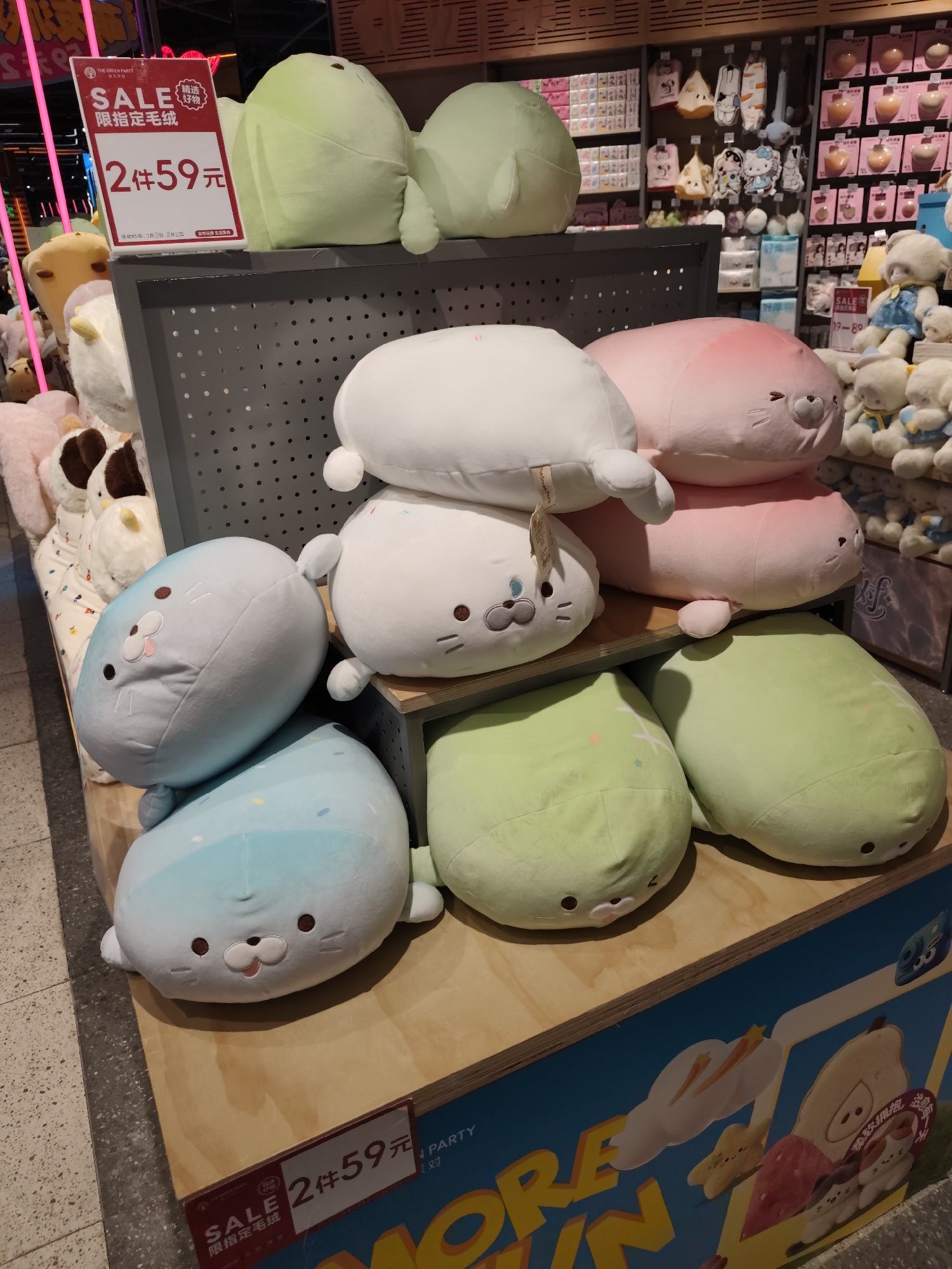 This image depicts a retail display of stuffed animals, possibly in a store located in Daqing, China. The stuffed animals are primarily seal-shaped and come in various colors, including light green, pink, white, and blue. They are stacked on wooden shelves and display stands. In the foreground, a red and white sale sign is visible, advertising "SALE" with Chinese characters indicating a promotion for "limited quantity fur balls" at a price of "2 items for 59 yuan". The background shows shelves stocked with other merchandise, some of which appear to be packaged goods. The lighting suggests an indoor retail environment. No people are clearly visible in the image.