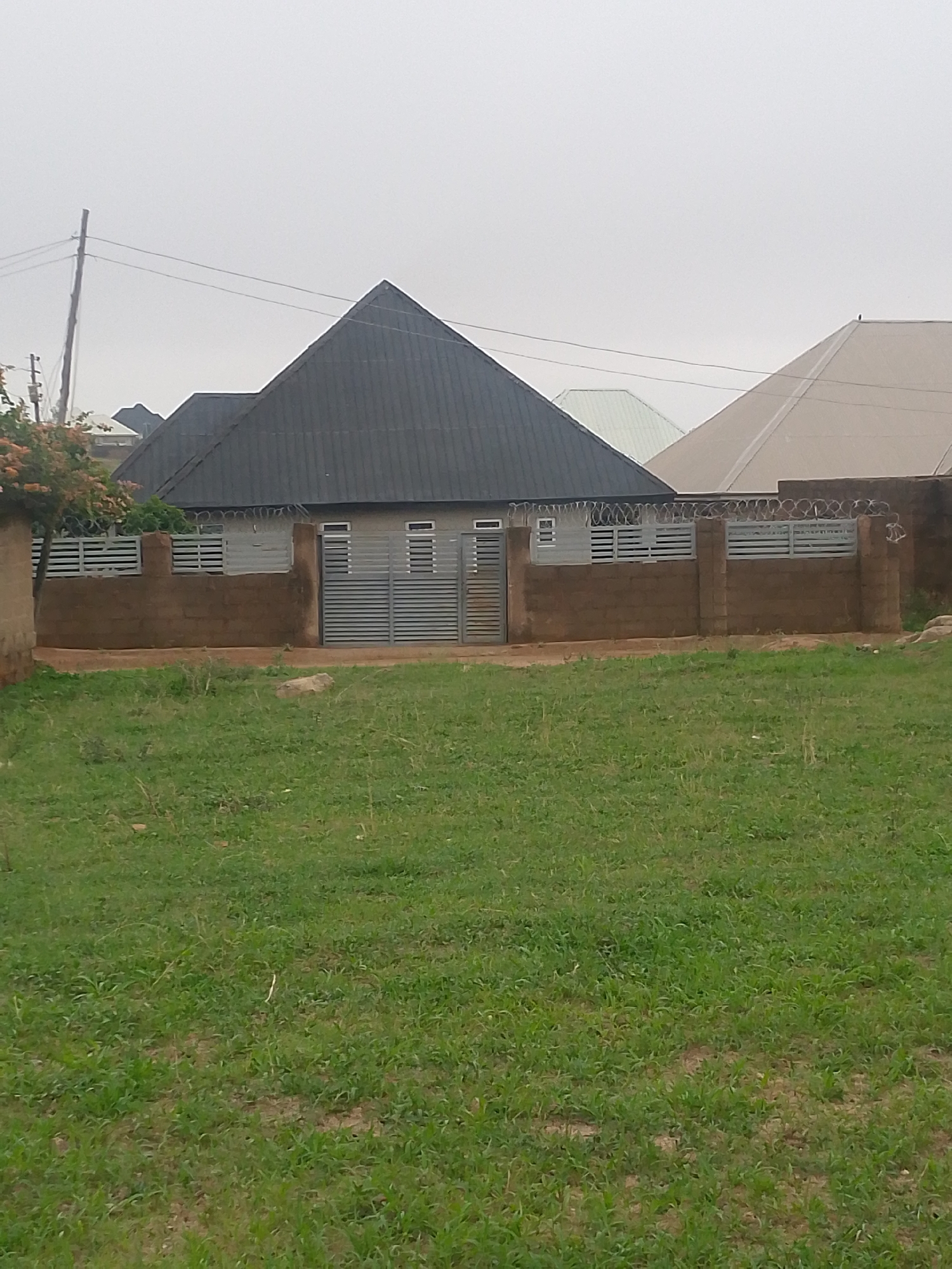 The image shows a house located in Barikin Gidar Kanat, Nigeria. The house has a dark grey, pyramid-shaped roof and a brown concrete block fence with horizontal metal slats. The gate is made of similar metal slats. Razor wire is visible on top of the fence. A grassy lawn extends from the fence towards the foreground. Other houses with lighter-colored roofs are visible in the background. The sky is overcast. A utility pole with wires is visible to the left of the house. There is a tree at the left edge of the image.