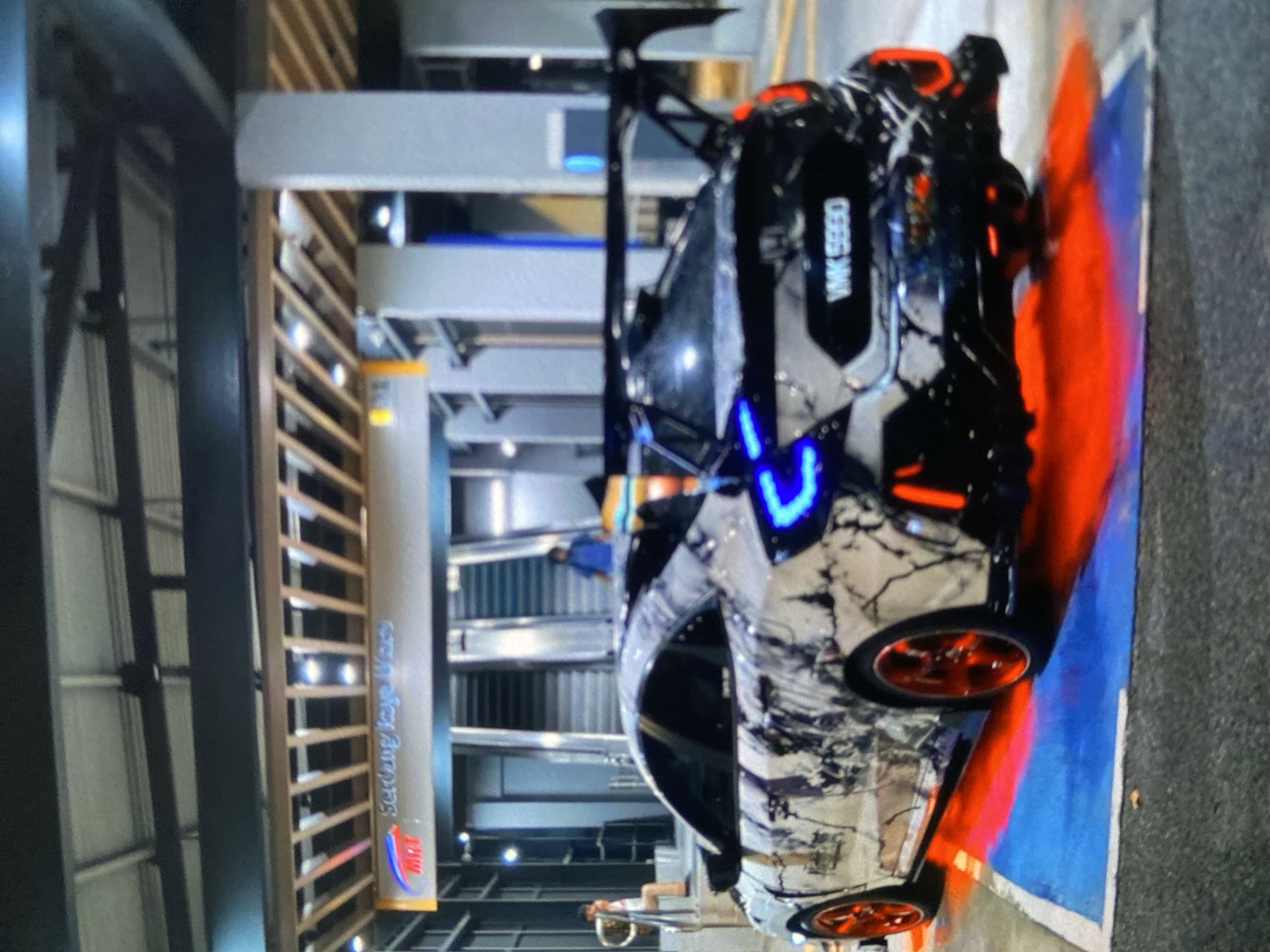 A highly modified, possibly custom, car is parked indoors, adjacent to a service structure with the words "Serdang Jaya Ware" on it. The car is predominantly white with a complex black pattern. It has orange rims, a large rear spoiler, and blue LED accents on the front. The license plate reads "VMK 5950". The car is parked next to a red and blue painted section of the pavement. Two people are faintly visible near the car, but their actions and details are unclear due to image quality and distance. The environment includes what appears to be a warehouse or indoor parking facility with metal construction elements. The location in the city of Lhokseumawe, Indonesia is not confirmed from the image, but the "Serdang Jaya Ware" signage could provide clues.