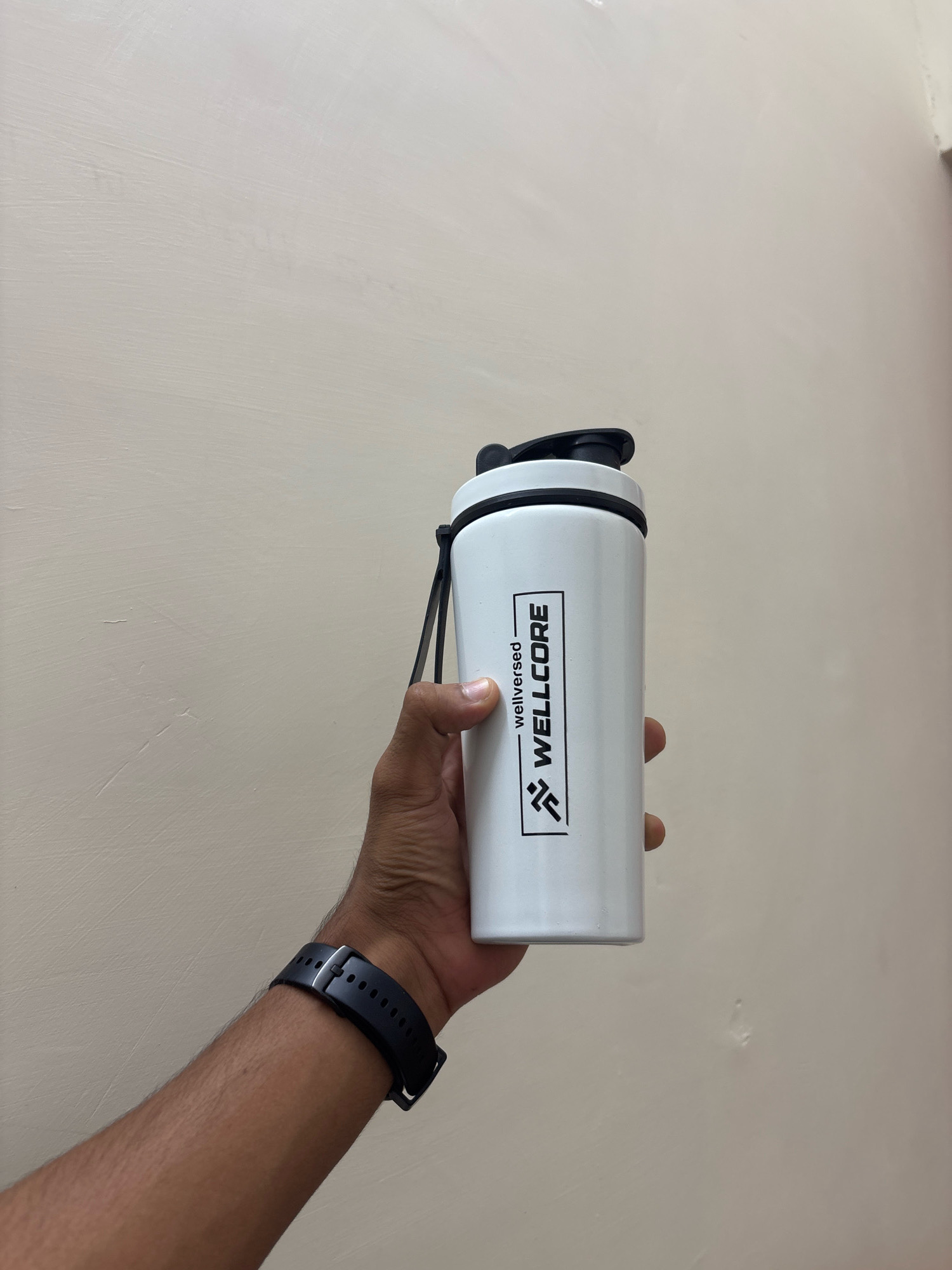 In an indoor, close-up shot, a person's right hand and forearm are visible, holding a white metal shaker bottle against a plain, beige wall. The person is wearing a black digital watch with a segmented band on their left wrist. The shaker bottle, which is white with a black lid and a black strap, features a black rectangular logo containing the text "wellversed WELLCORE" in white and a stylized graphic of two figures running. The background wall has a textured, matte finish and appears to be slightly uneven. The lighting is even and diffuse.