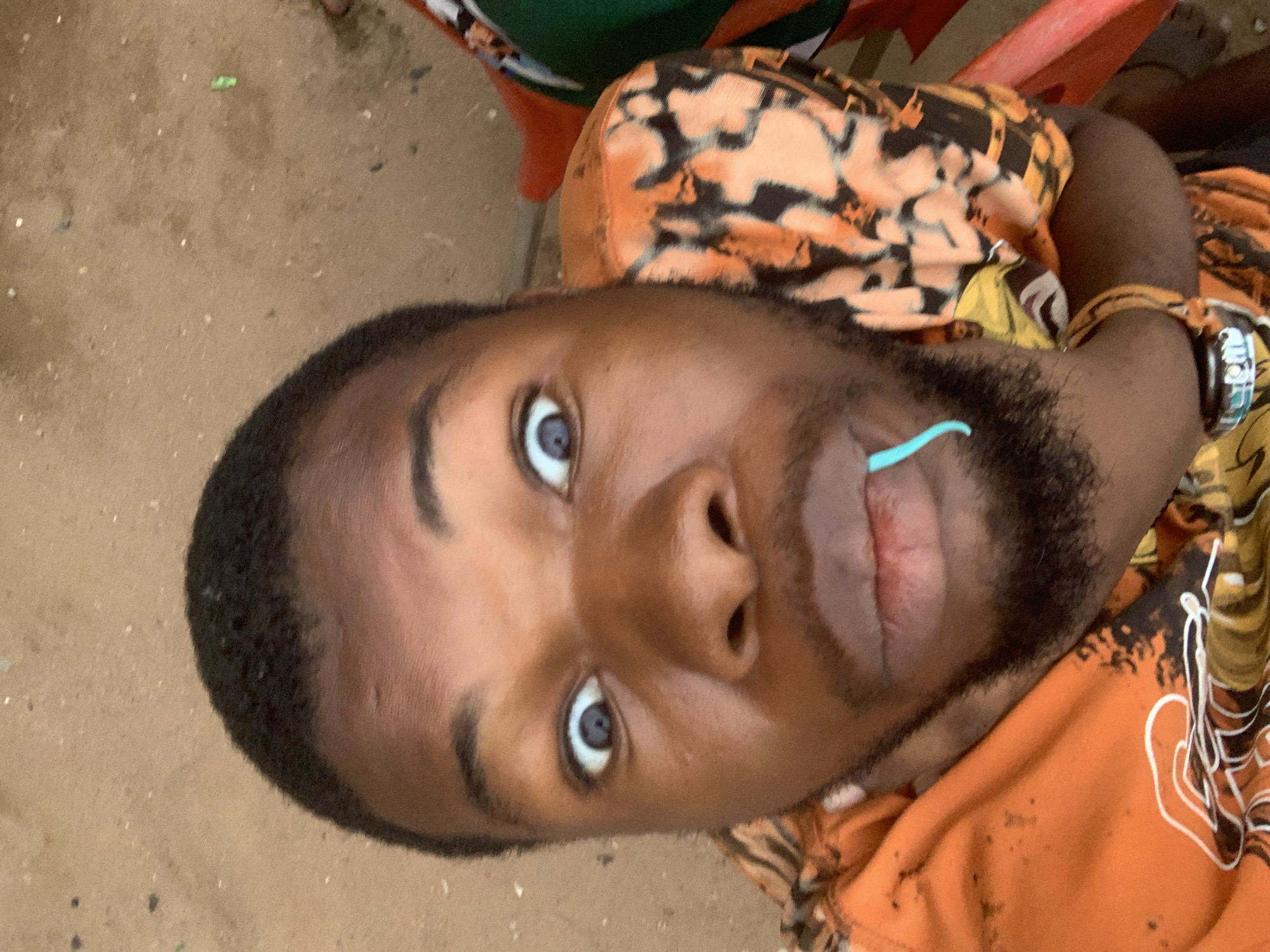 This rotated image captures a close-up portrait of a young Black man, likely in Onitsha, Nigeria. He has a dark complexion, short dark hair, and a neatly trimmed beard. His striking light blue eyes look directly towards the viewer. A light blue, curved object, possibly a candy stick or a small straw, is held between his slightly parted lips.
He is wearing an orange shirt with an abstract black pattern, reminiscent of animal print. A watch with a metallic and blue-toned band is visible on his right wrist. The man appears to be leaning back or lying down, with the photo taken from a high angle looking down at him.
The background consists primarily of a sandy or dusty light brown ground, suggesting an outdoor or semi-outdoor environment. Small specks and debris are scattered across the ground. In the upper part of the frame, hints of red and green objects, possibly parts of a chair or other furniture, can be seen. The lighting is soft and natural, indicating daytime. There is no visible text within the image. His expression is calm and direct, with a hint of playfulness from the object in his mouth.