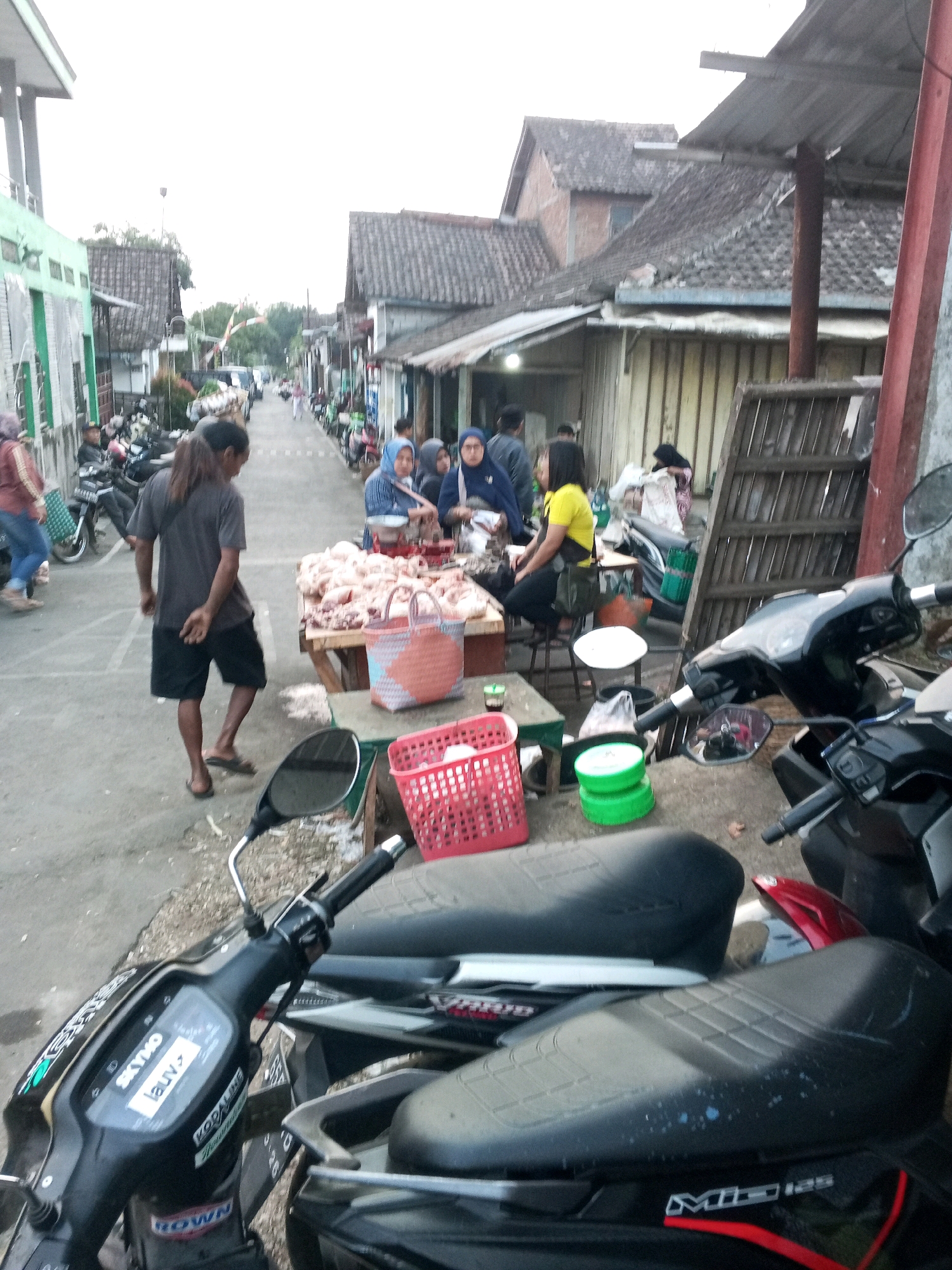 In Karanggede, Indonesia, a street market is set up along a narrow road. Several people are gathered around tables displaying raw chicken for sale. A man with long hair, wearing a grey t-shirt and black shorts, stands near the market stall, facing away from the camera. A woman in a yellow shirt and black pants sits at a stall, while other individuals, some wearing hijabs, are also present. Motorcycles are parked along the sides of the road, with two of them prominently featured in the foreground. The buildings lining the street have tiled roofs and wooden walls. The scene appears to be an outdoor marketplace during daylight hours, with a general atmosphere of commerce and local activity.