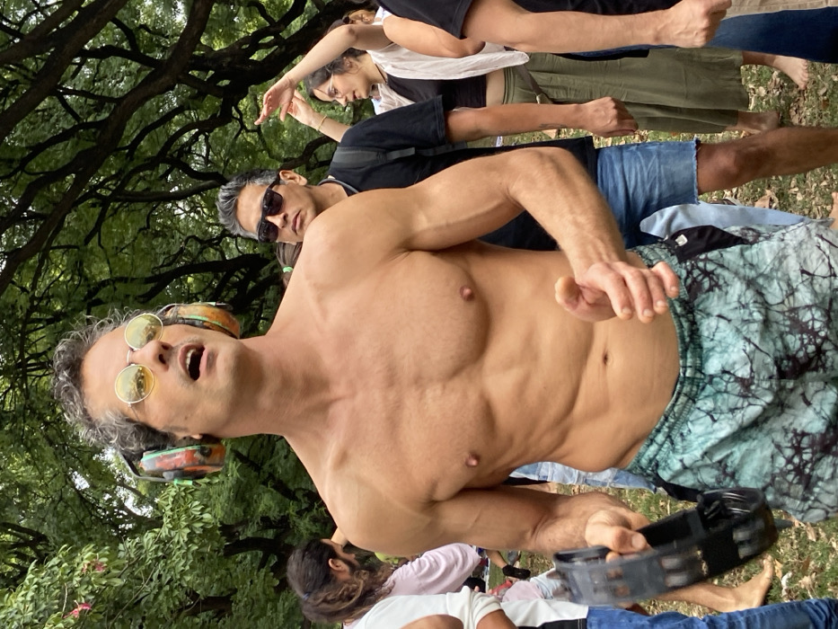 The image depicts an outdoor gathering in a park within the city of Buenos Aires, Argentina, likely in the Palermo neighborhood, characterized by a dense canopy of mature trees with dark branches and green foliage. The central figure is a shirtless, muscular man with light skin and short, curly gray hair. He wears small, round, yellow-tinted sunglasses with thin silver frames and multi-colored over-ear headphones (orange, green, black). His mouth is open, suggesting vocalization or singing, as he looks upward and slightly to his right. In his left hand, he holds a black tambourine; his right hand is open with fingers slightly curled. He wears turquoise and black patterned shorts. Behind him, to his right, a man with gray hair, dark sunglasses, and a black t-shirt stands, looking forward. Further in the background, to the right of the central figure, a woman wearing glasses, a light-colored cardigan over a dark top, and a long olive-green skirt has both hands raised with fingers splayed, indicating movement or dance. Other participants are partially visible in the blurred foreground and background. The ground consists of grass and sparse vegetation.