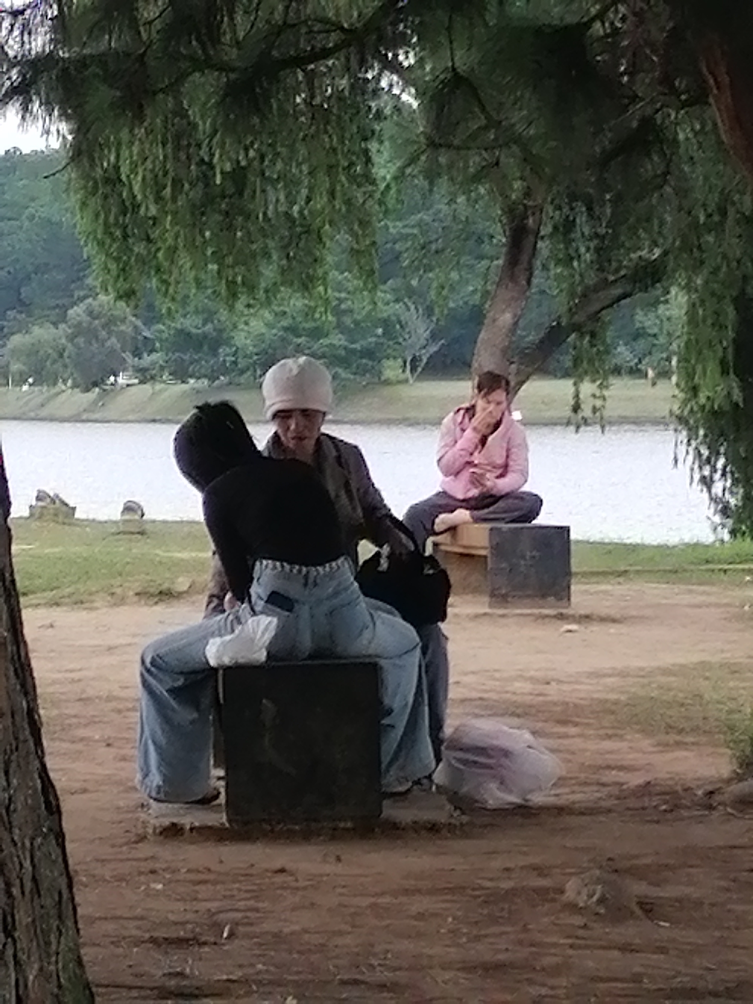Two individuals are seated near a body of water, possibly a lake, in Da Lat, Vietnam. The person in the foreground, facing away from the camera, is wearing a black top and light blue jeans, and is seated on a dark, cubic object. This individual has a white piece of cloth tucked into the back of their jeans. Another person, wearing a light pink jacket and dark pants, is seated cross-legged on a wooden bench in the background, looking towards the camera and touching their face. The environment appears to be a park with trees, grass, and a path.