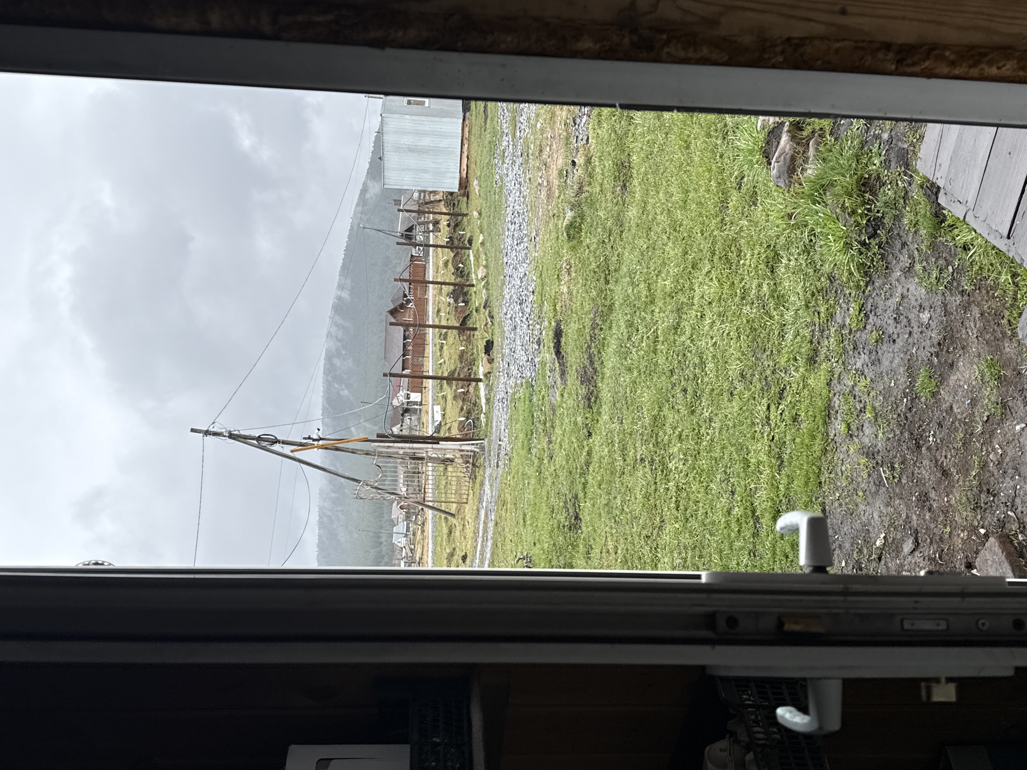 The image shows an exterior view from inside a building, potentially through an open window. The scene outside depicts a rural or semi-rural environment, possibly in or near Tyulyuk, Russia. The landscape features a grassy area leading to a gravel or rocky section. In the background, there are some buildings, wooden structures or fences, and a utility pole with wires. A mountain or hill is visible in the distance, shrouded in mist or low clouds, suggesting overcast or rainy weather. The immediate foreground includes parts of the window frame and what appear to be interior elements of the building. The ground near the window appears muddy.