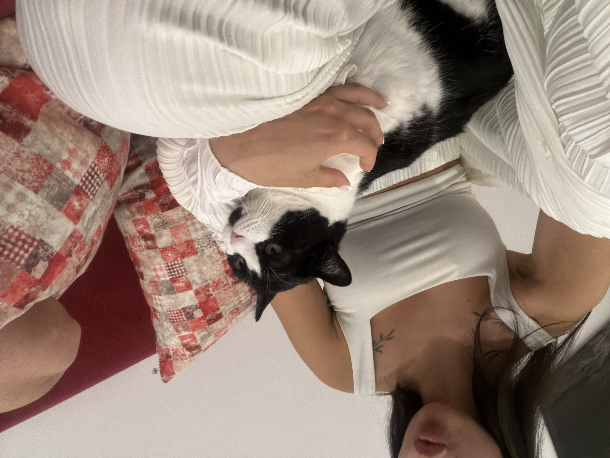 A person, dressed in white clothing, is sitting on a red sofa with patterned pillows and holding a black and white cat. The person's face is not fully visible, but their lips are slightly parted as if about to speak. The cat is looking to the right with its eyes open. The scene appears to be indoors, possibly in a living room. There is no indication of the city of Burgas, Bulgaria, in the image.