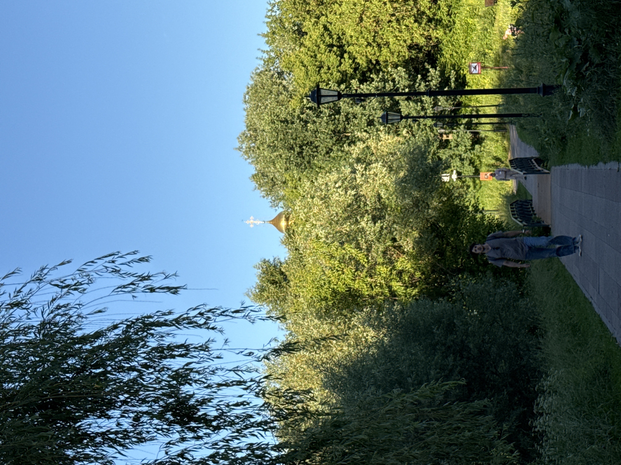 The image depicts a sunlit pathway in a park, likely during the late afternoon, given the golden hue of the light. The pathway, made of light-colored paving stones, curves gently through lush green foliage. On the left, a man in a grey t-shirt and blue jeans walks towards the viewer, carrying a backpack. He is positioned on the pathway and appears to be mid-stride.
In the background, several black lampposts line the pathway, with ornate tops and single bulbs. Beyond the path, a dense array of green trees creates a vibrant natural backdrop. Peeking through the canopy of leaves, a golden dome of a church, topped with a cross, is visible. Further down the path, a smaller bridge spans a low point, and a person in lighter-colored clothing can be seen walking away from the viewer. The sky is a clear, bright blue. The scene evokes a sense of tranquility and natural beauty within an urban setting. This scene is likely within the grounds of Kolomenskoye in Moscow, Russia, known for its parkland, historical architecture, and the presence of the Church of Ascension.
